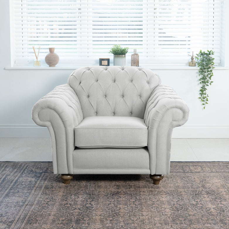 A Chesterfield Armchair in Warm Grey takes center stage, its tufted elegance handcrafted in England. Resting on a patterned rug and framed by potted plants with blinds in the background, it embodies traditional British design and timeless appeal.