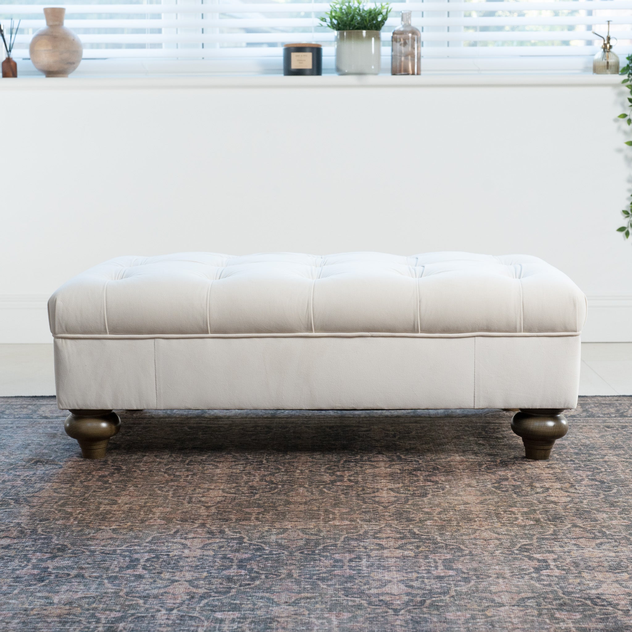 The Chesterfield Velvet Footstool in Cream, with its classic tufted design, enhances the patterned rug while framed by lush plants and elegant blinds.