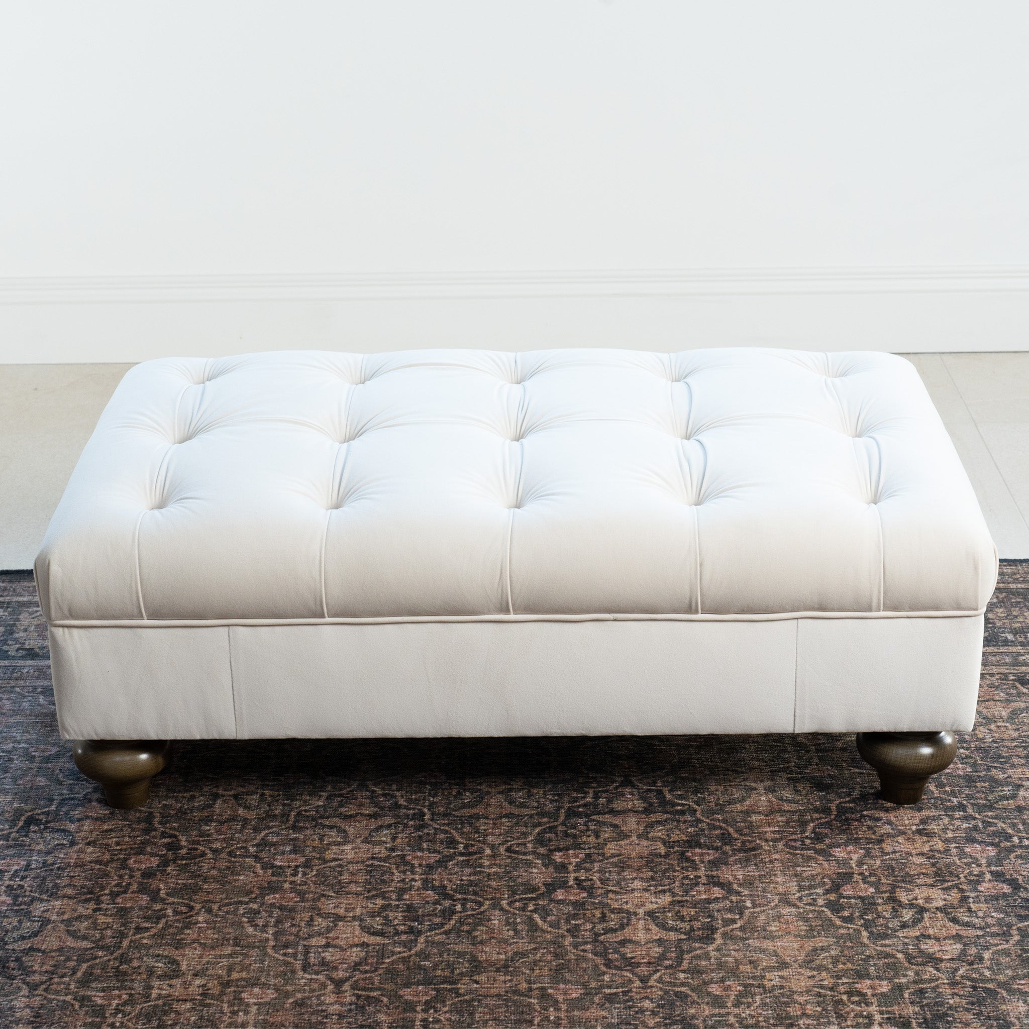 The Chesterfield Velvet Footstool in Cream, featuring tufted design and dark wooden legs, sits elegantly on a patterned rug in a minimalist room.