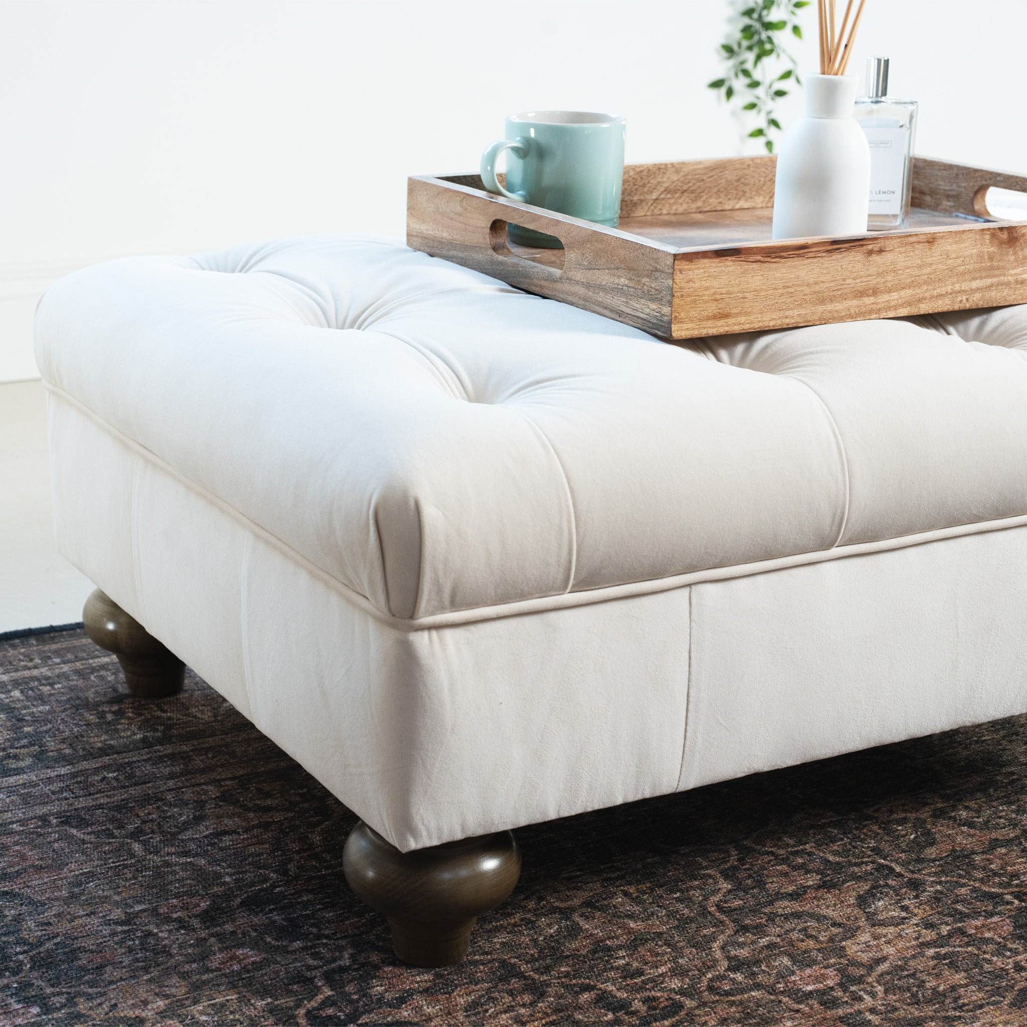 The Chesterfield Velvet Footstool in Cream, with tufted design, supports a wooden tray holding a mug and decor, elegantly placed on a patterned rug.