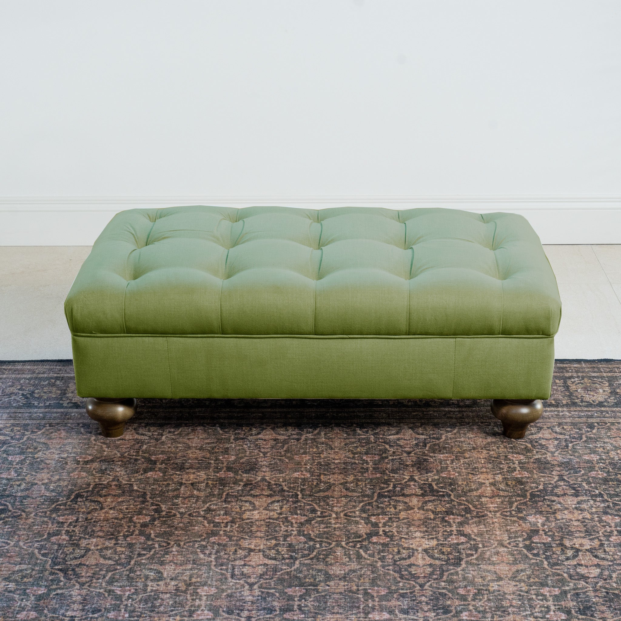 An Olive Chesterfield Footstool with wooden legs sits gracefully on a patterned rug against a light wall, showcasing classic design elements that echo timeless British style.