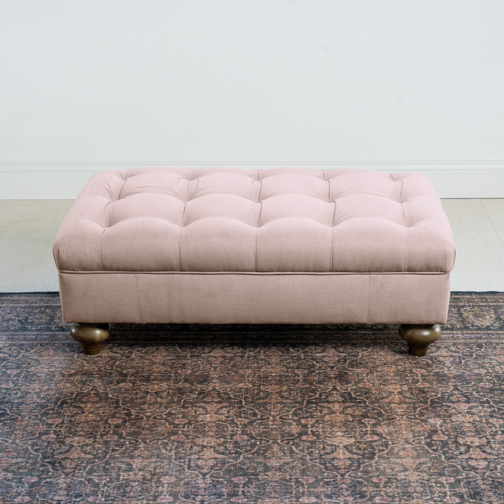 A Chesterfield Footstool in Dusky Pink is elegantly placed on a dark patterned rug in a minimalist room.