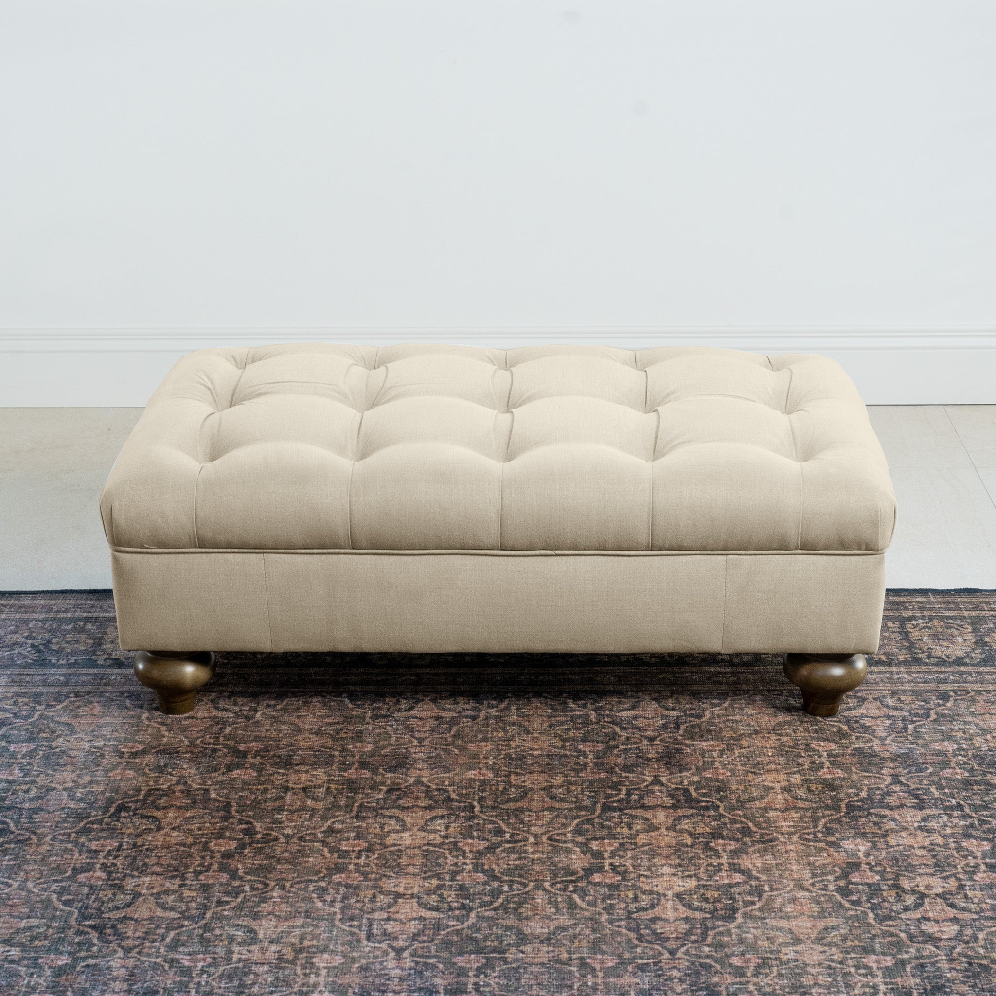 The Chesterfield Footstool in Beige, featuring artisanal upholstery and round wooden legs, exudes classic British elegance while resting gracefully on a patterned rug.