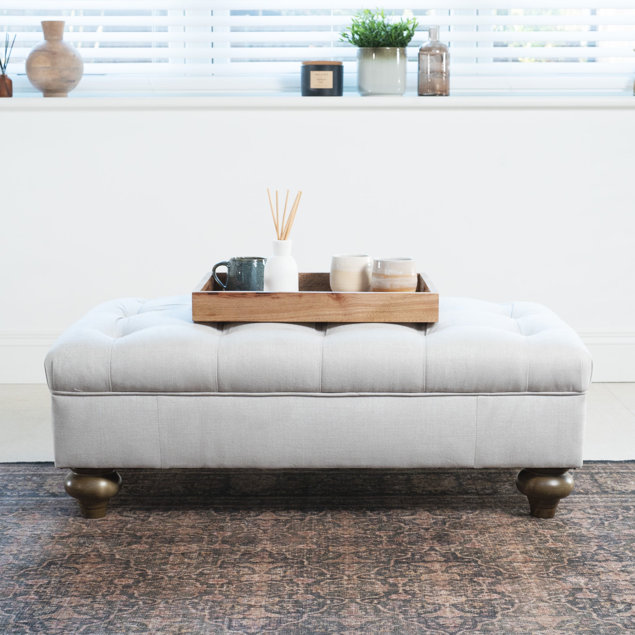 Warm grey Chesterfield Footstool with tufting holds a wooden tray and decor in a bright room with a rug and plants, featuring traditional artisan upholstery.