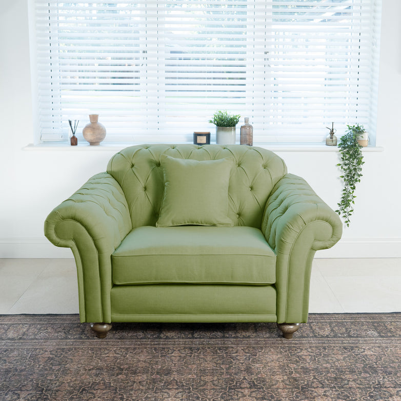 A Chesterfield Loveseat in Olive, featuring deep-button tufting, complements the green upholstered armchair with a cushion in the bright room. Both pieces mingle harmoniously in front of a window with blinds and decorative items, creating an inviting and stylish space.