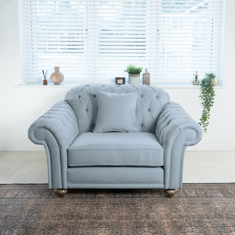 A Chesterfield Loveseat in Sky Blue with a cushion sits gracefully in front of a window with blinds, set on a patterned rug, enhancing the sustainable furniture theme of the room.