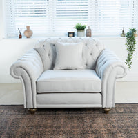 A Chesterfield Loveseat in Warm Grey with a cushion is placed by a window with blinds, accompanied by decorative items and a handcrafted sofa.