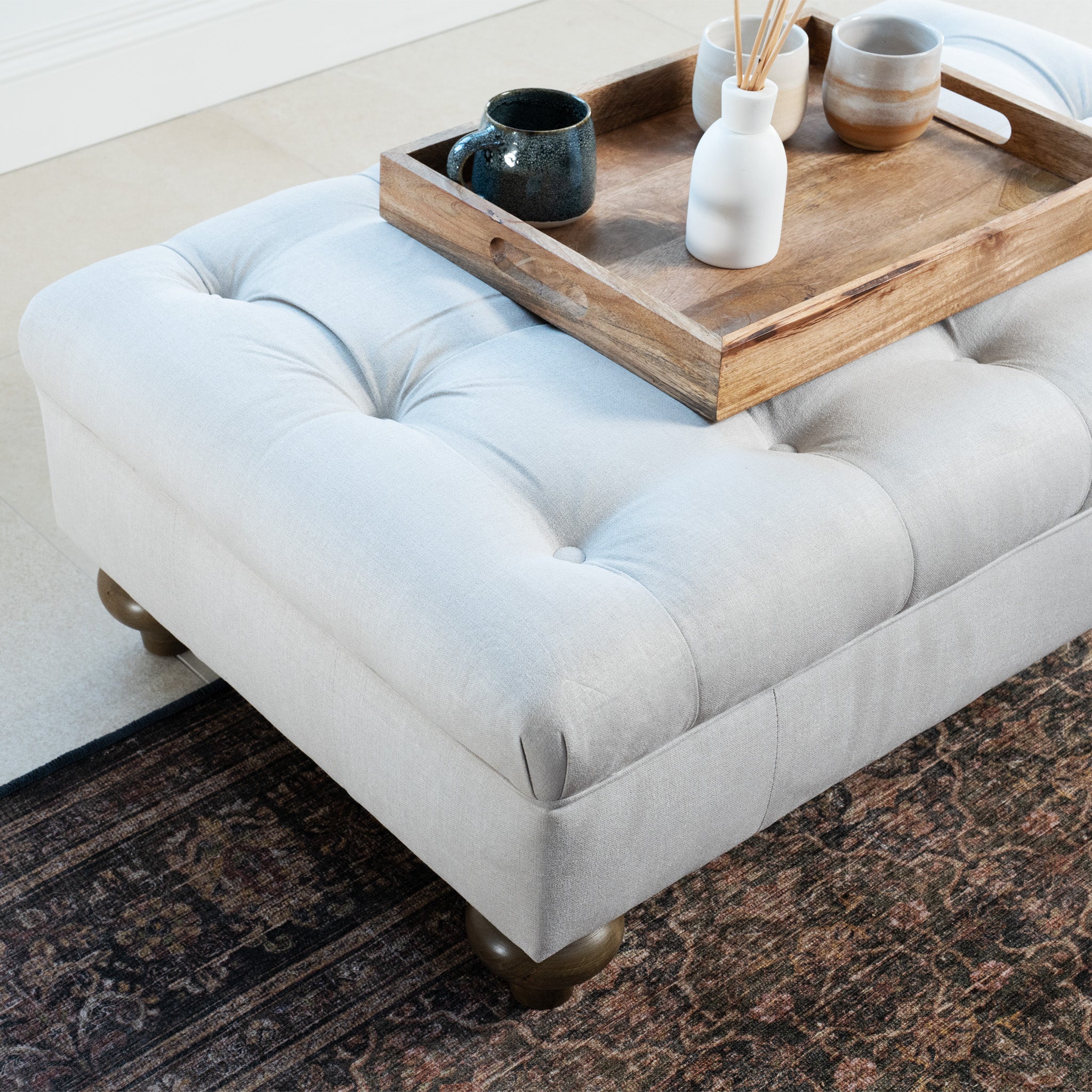 A wooden tray with mugs and a vase sits elegantly on the Chesterfield Footstool in Warm Grey beside a patterned rug, highlighting traditional artisan upholstery.