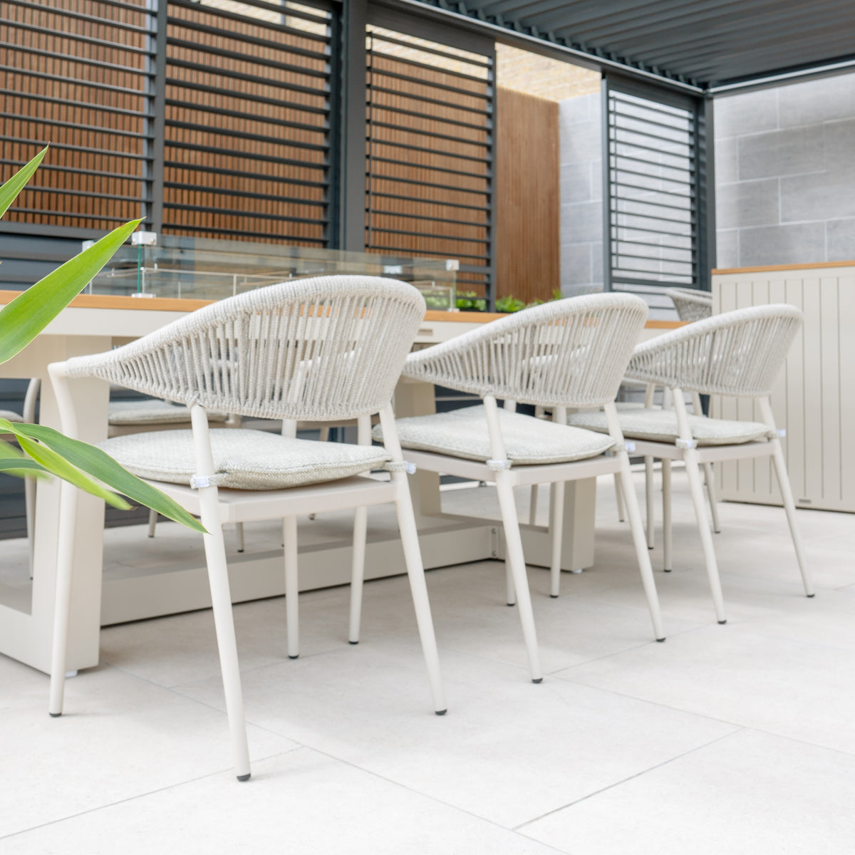 The white cushioned chairs from the Cloverly 8 Seat Rectangular Teak Firepit Dining Set in Latte are arranged around a table in a modern outdoor patio seating area, with plants in the foreground. The UV-resistant chairs feature a hydrophobic rope weave for added durability and style.