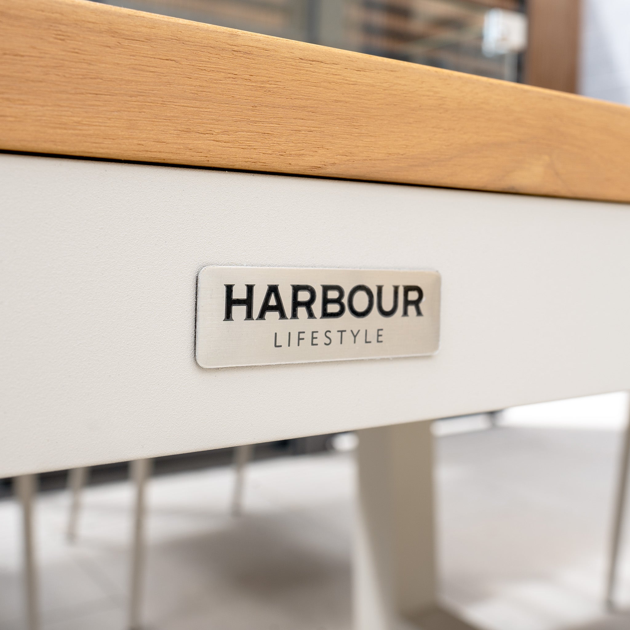 Close-up of a metal label on a piece of furniture from the Cloverly 8 Seat Rectangular Teak Firepit Dining Set in Latte that reads "Harbour Lifestyle." The table has a white base and wooden top.