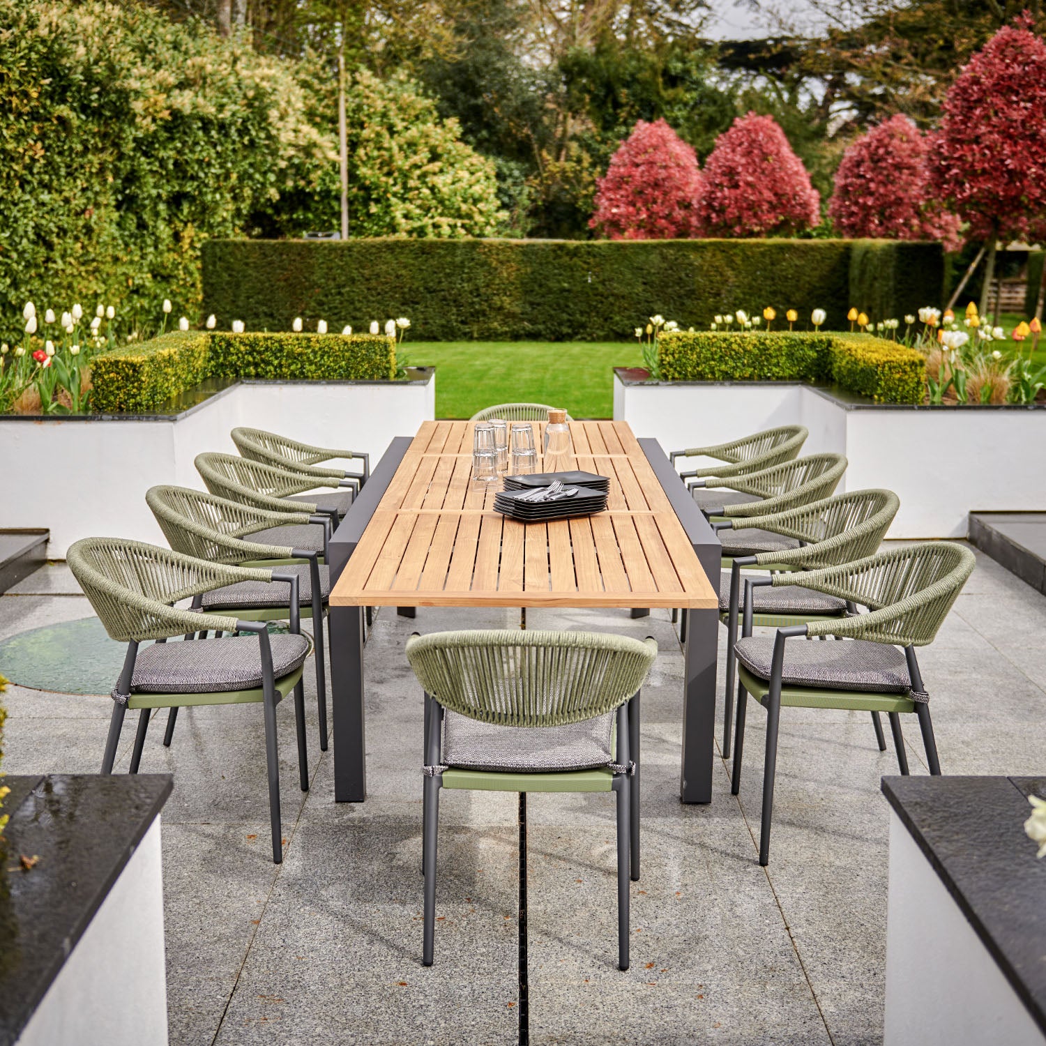 Cloverly 8 Seat Rectangular Extending Dining Set with Teak Table in Olive Green, showcased on a patio surrounded by hedges and blooming plants. The weather-proof furniture ensures long-lasting enjoyment for outdoor gatherings.