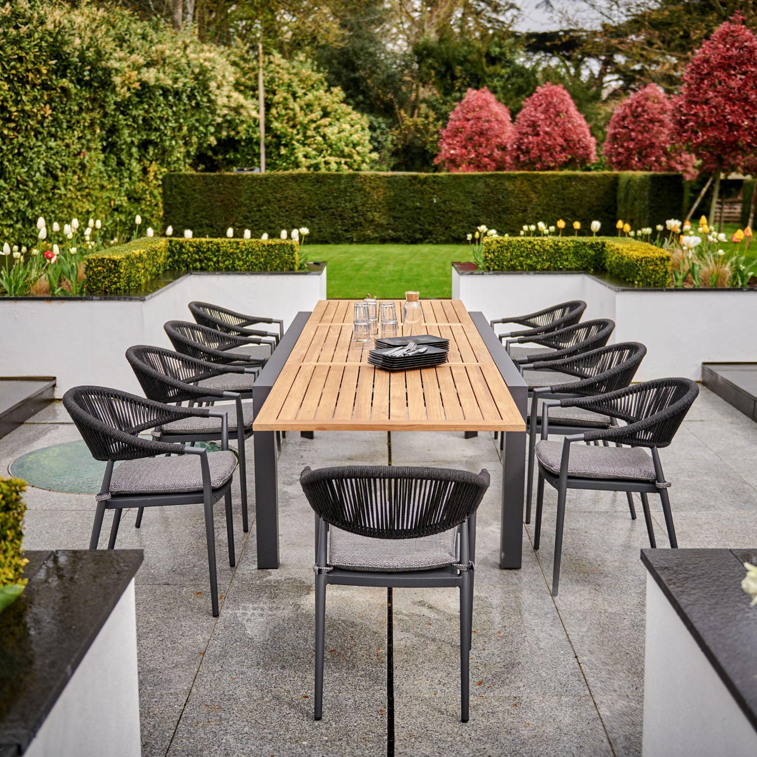 Cloverly 8 Seat Rectangular Extending Dining Set with a teak table in charcoal and black woven chairs, surrounded by greenery, flowers, and manicured hedges.