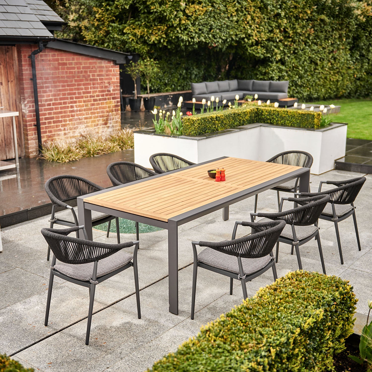 Cloverly 8 Seat Rectangular Extending Dining Set with a charcoal teak table and woven chairs, placed on a patio surrounded by greenery with a brick building in the background.