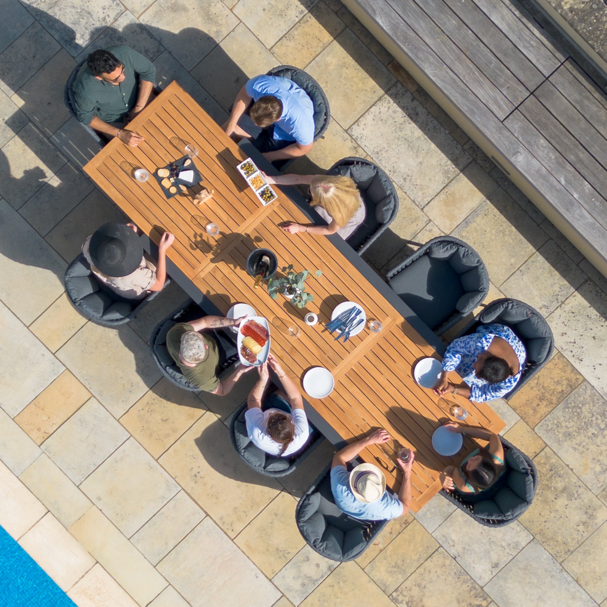 Aerial view of people gathered around a Monterrey 10 Seat Rope Extending Teak Dining Set in Grey adorned with food and drinks near a pool, seated comfortably in Monterrey Chairs.