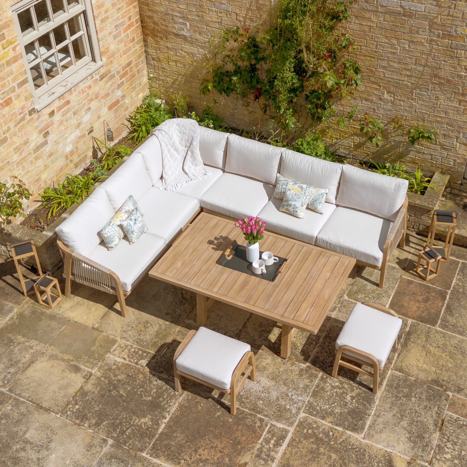 Aerial view of an outdoor patio featuring the Quay Corner Dining Set in Linen, showcasing a white cushioned L-shaped sofa, a wooden table, stools made from sustainably sourced acacia wood, and plants against brick walls. The water-resistant polyester fabric ensures durability in all weather conditions.