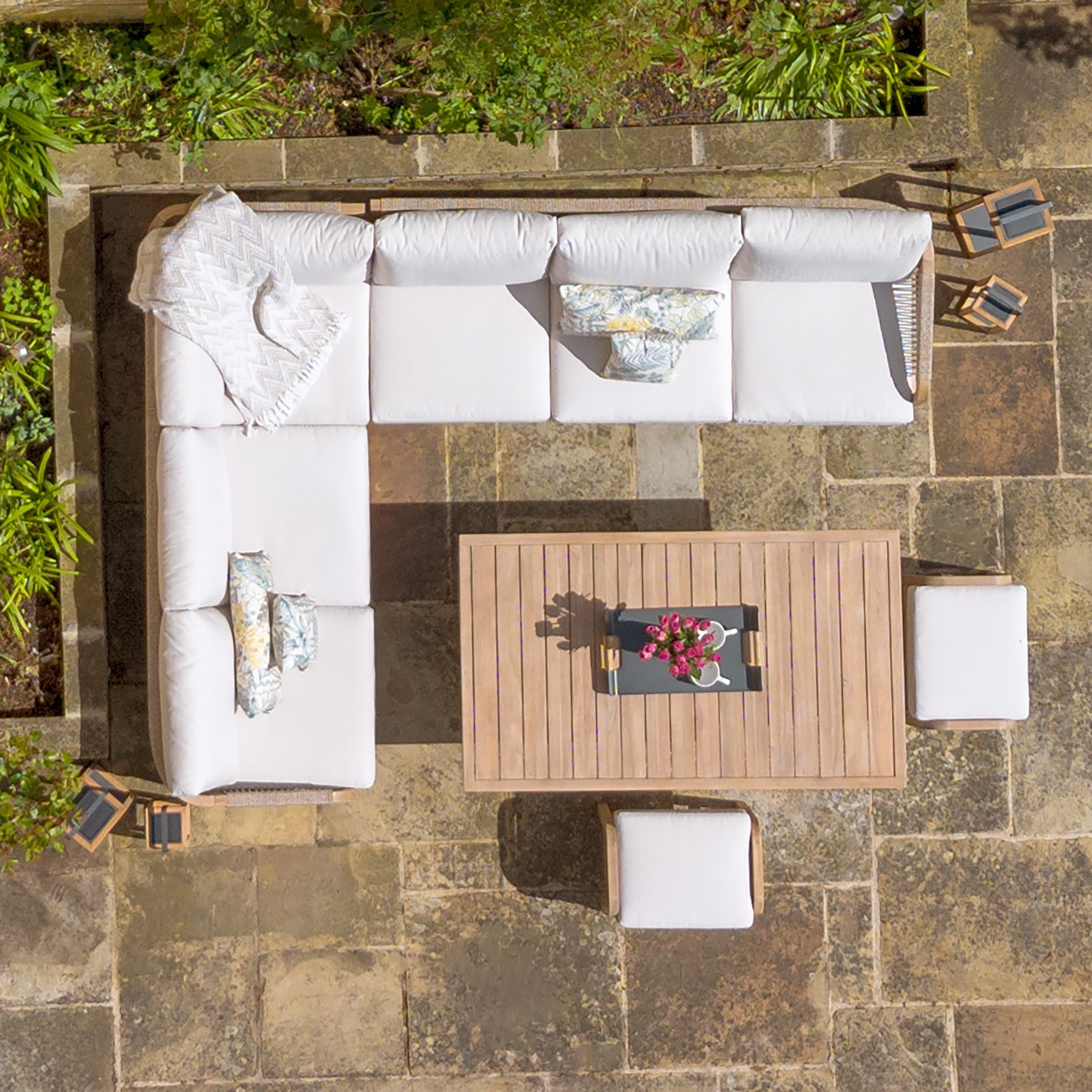 Aerial view of a patio featuring the Quay Corner Dining Set in Linen with an L-shaped white sofa, wooden table made from sustainably sourced acacia wood, cushioned stools with water-resistant polyester fabric, and outdoor decor on stone flooring.