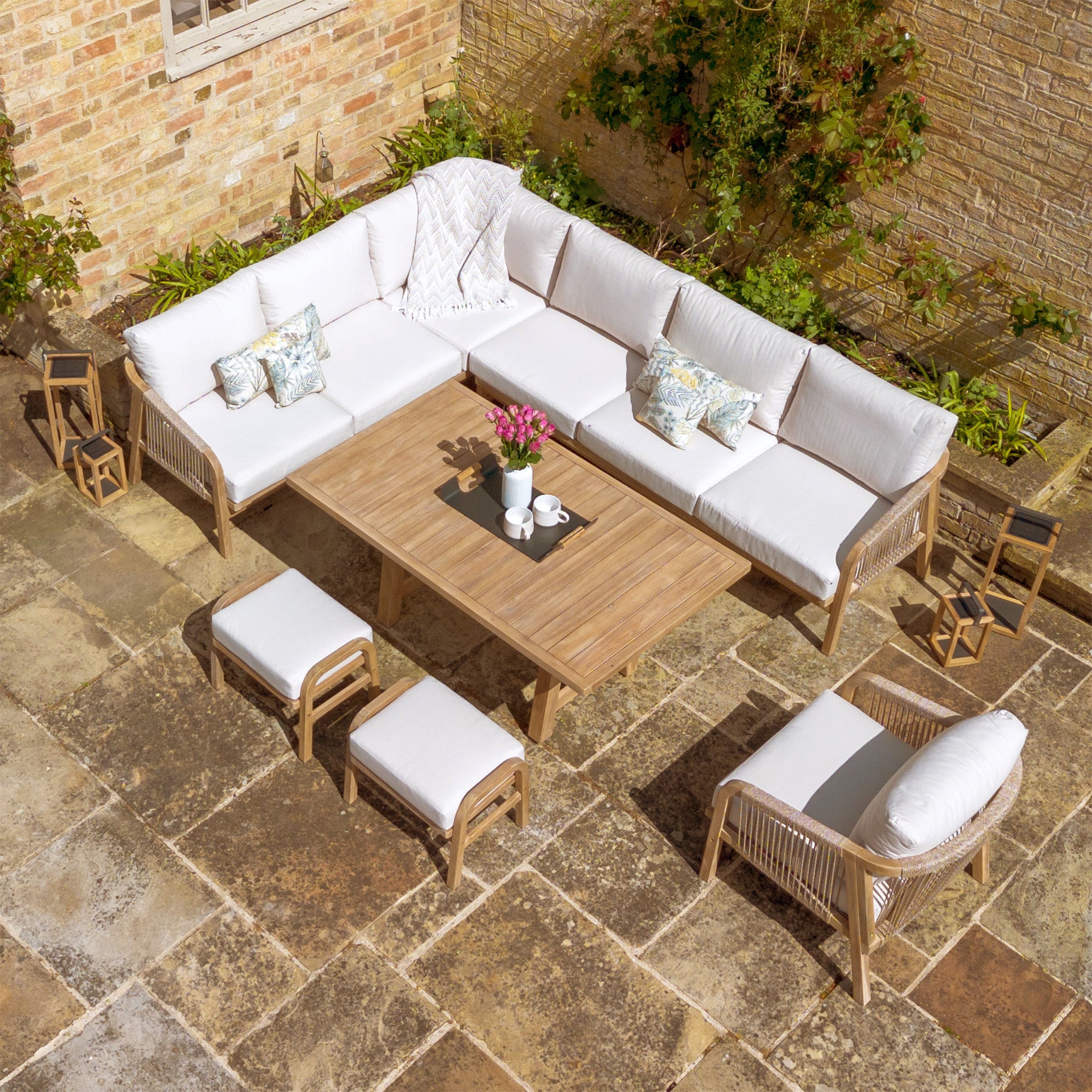 Outdoor seating setup featuring a Quay Corner Dining Set with Armchair in Linen, accompanied by stools on a stone patio—all crafted from sustainably sourced acacia wood.