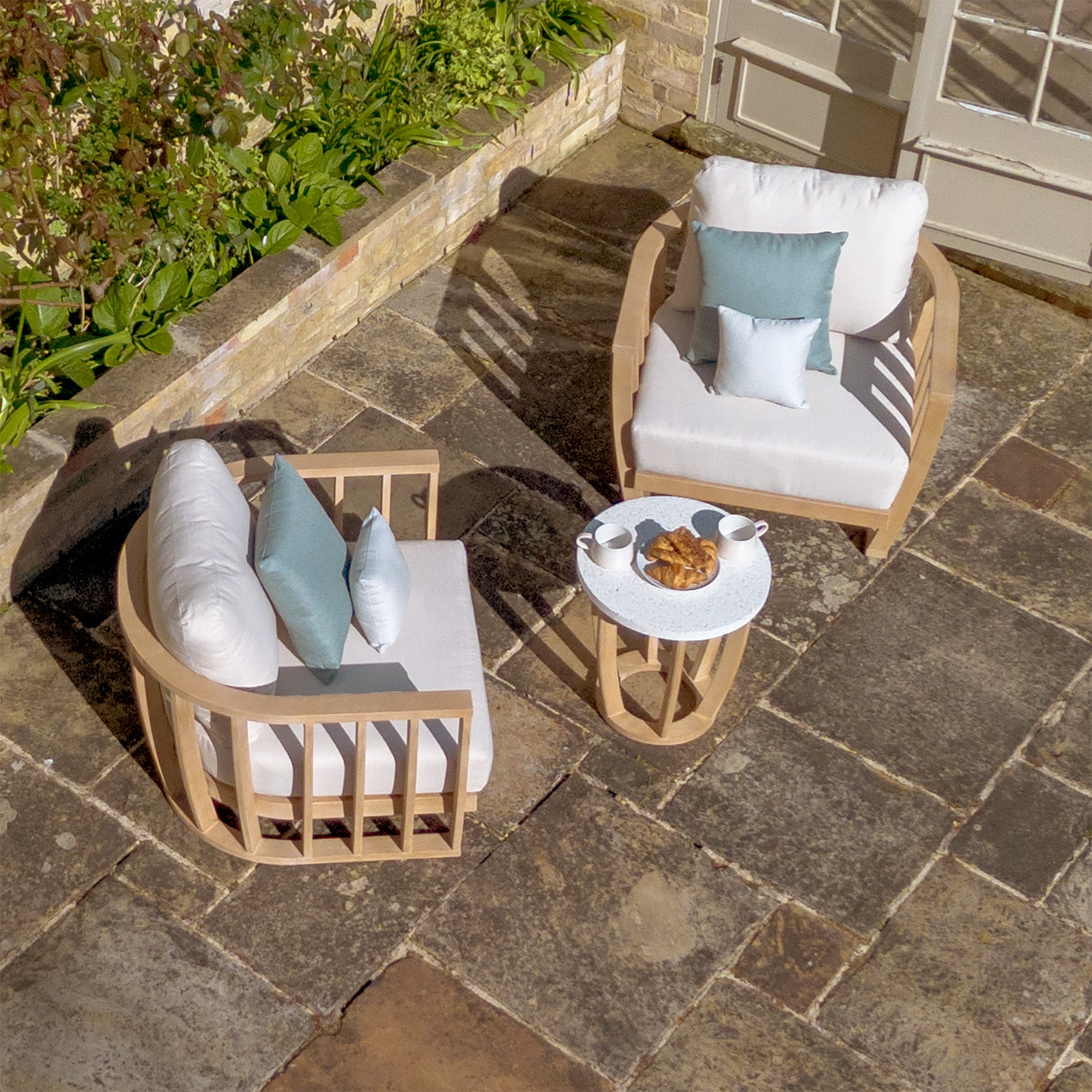 Two cushioned wooden chairs with deep plush cushions and pillows, part of the elegant Cape Lounge Set in Beige, are placed around a small table holding coffee cups and pastries on a stone patio near a garden.