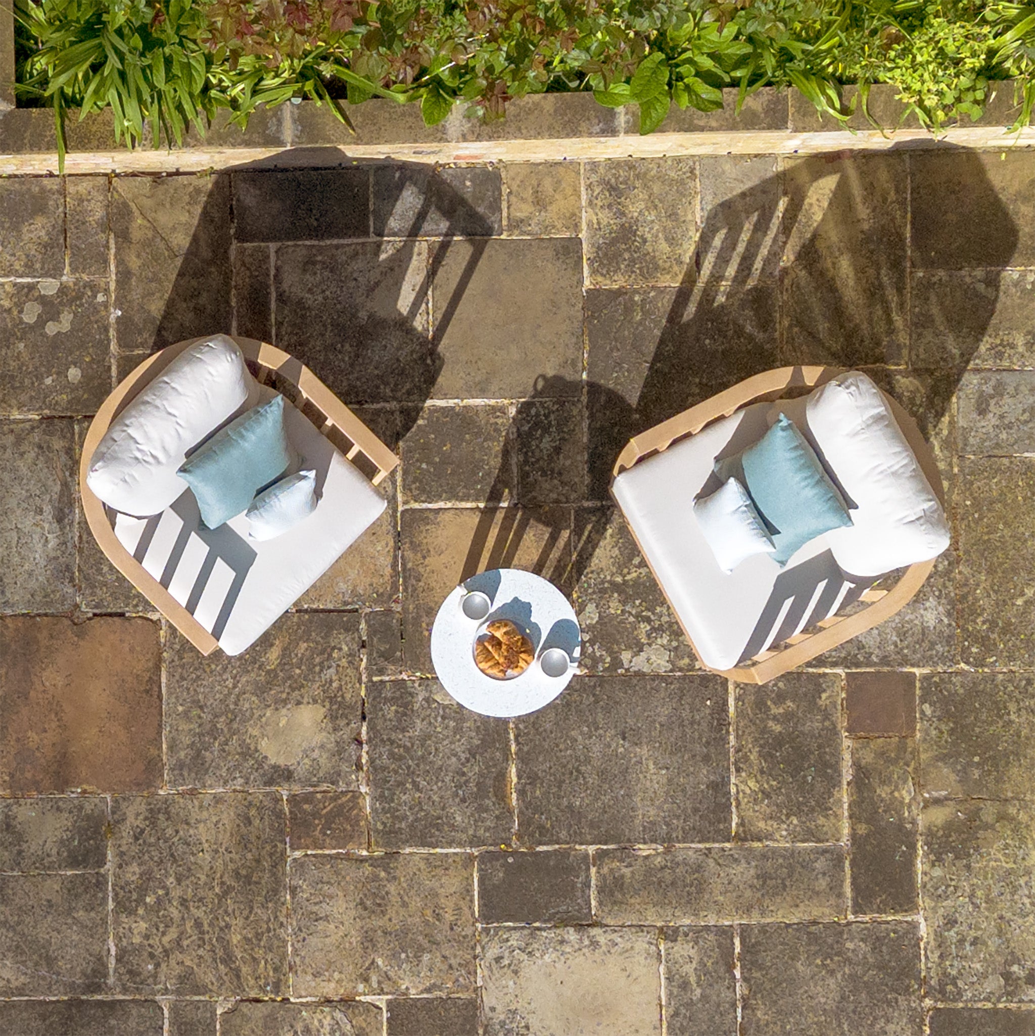 Two cushioned outdoor chairs from the Cape Lounge Set in Beige, featuring deep plush cushions and pillows, sit around a small table holding drinks and snacks on a stone patio.