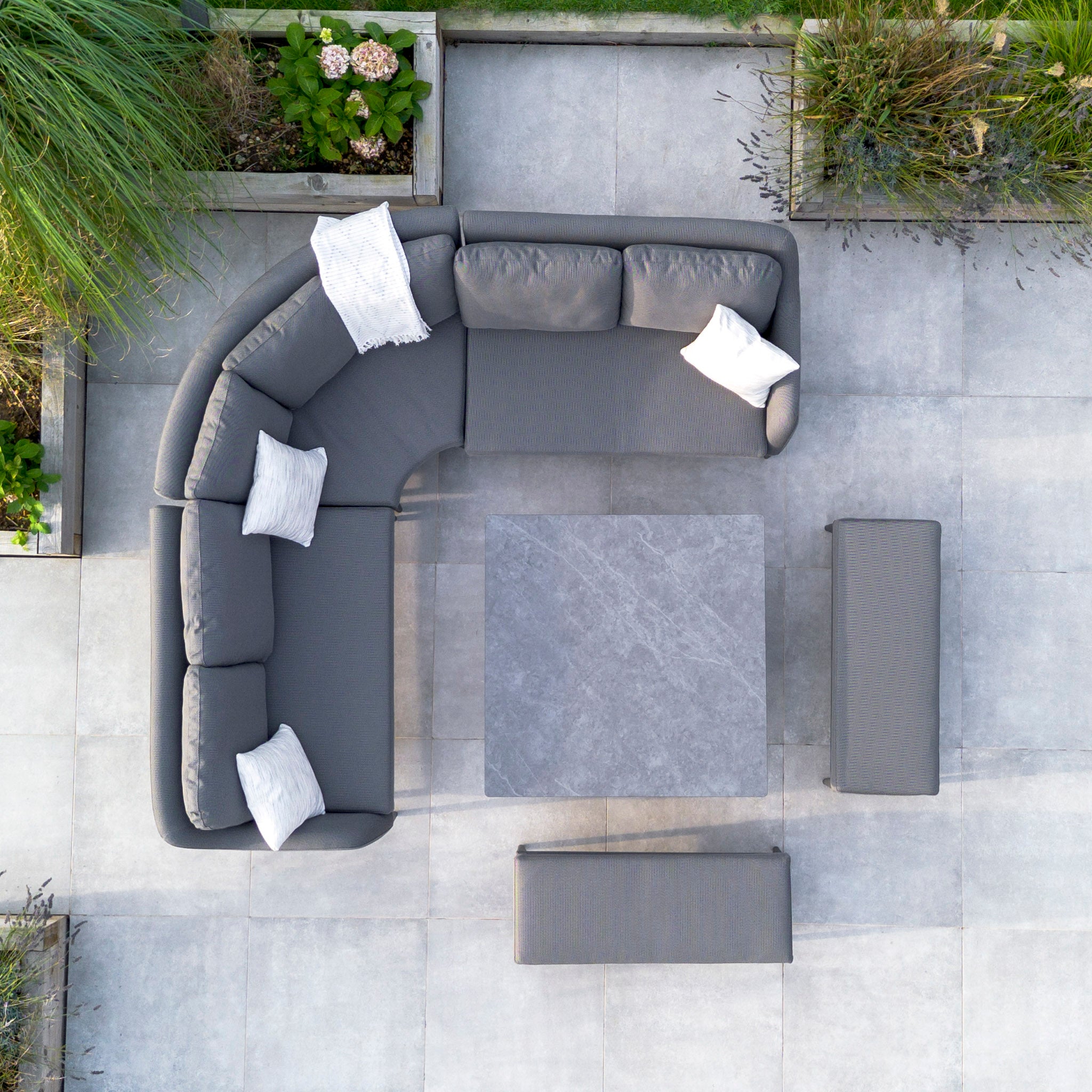 Aerial view of a modern outdoor patio featuring the Luna Deluxe Outdoor Fabric Square Corner Dining Set with Rising Table in Grey, complete with gray sofa, white pillows, and a square stone table in the center. This elegant arrangement showcases all-weather material for lasting durability and style.