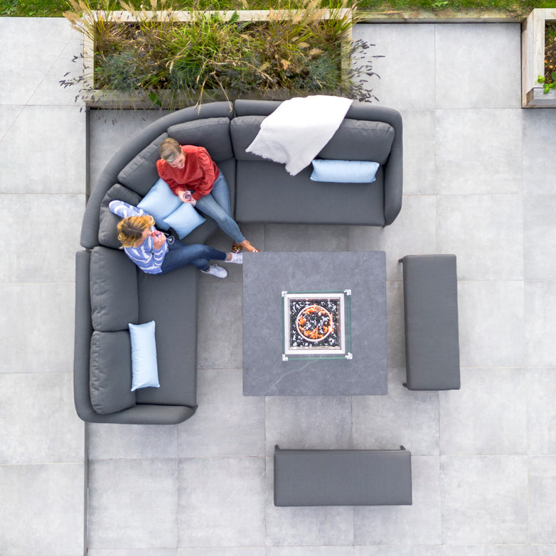 Two people sit on the Luna Deluxe Outdoor Fabric Square Corner Dining Set with Rising Firepit Table in Grey, engaging in conversation on a paved patio. Top-down view.