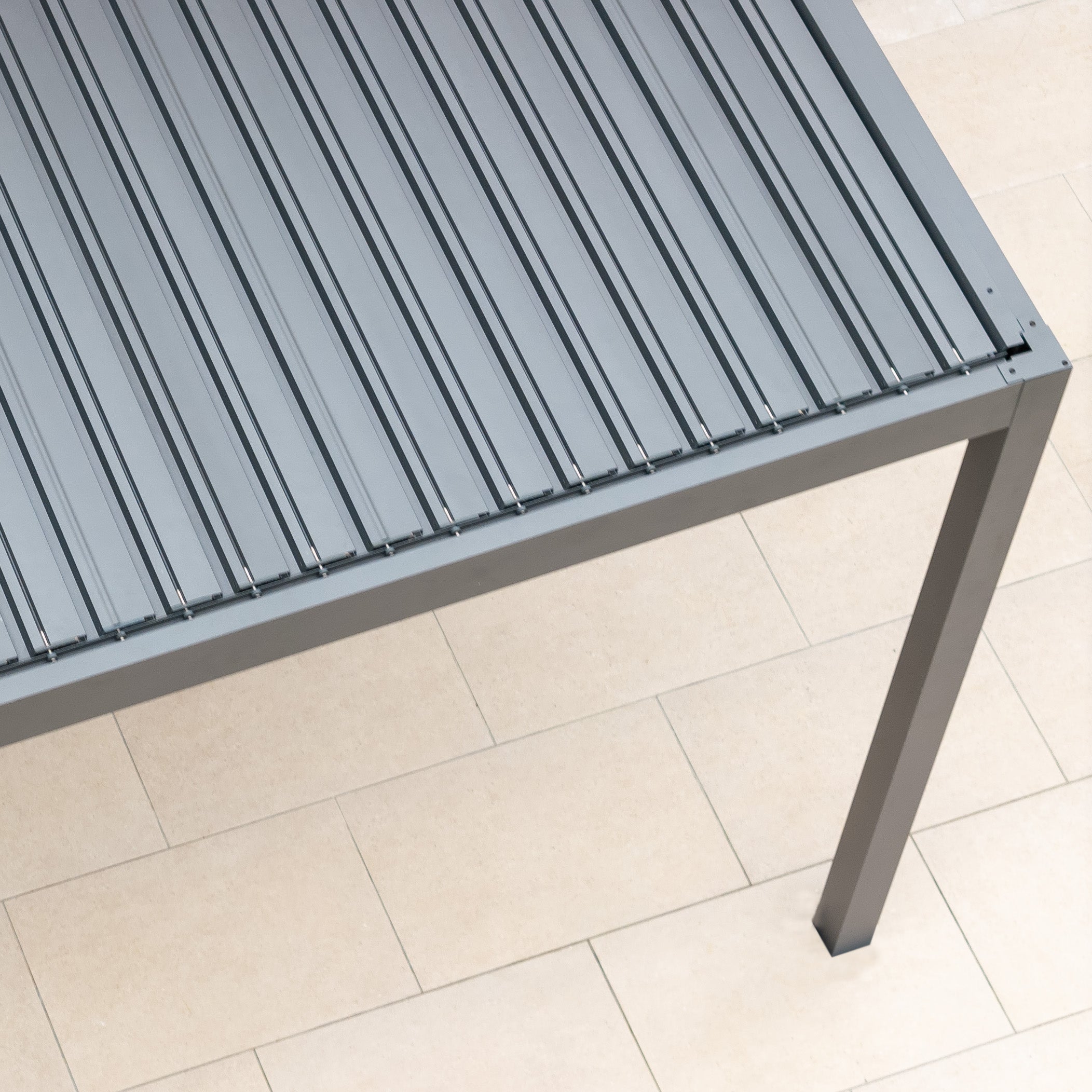 A grey slatted metal table with four legs is elegantly positioned on a beige tiled floor beneath the PergoSTET Deluxe Frame Only 3m x 4m Rectangular Pergola, featuring motorised LED louvres in Grey.