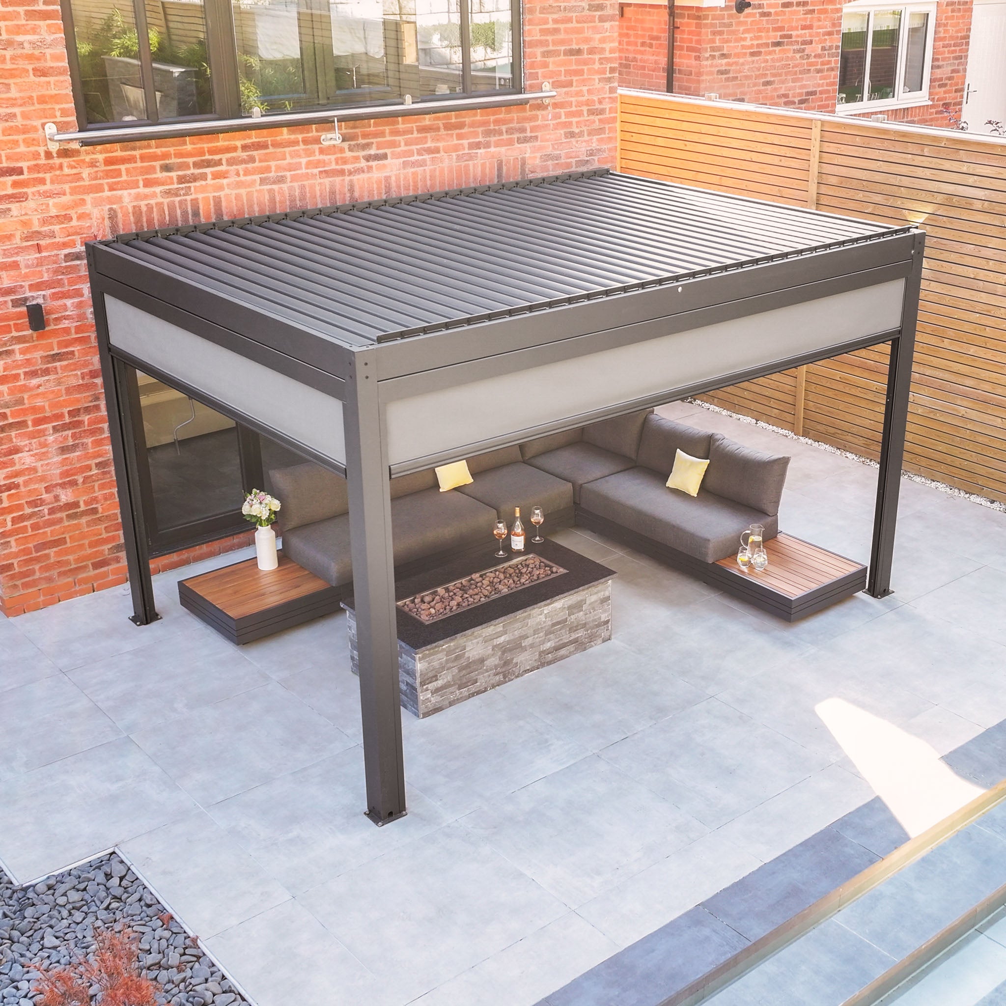 A modern outdoor patio featuring a PergoSTET Plus 3m x 4m Rectangular Pergola with Drop Sides and LED Lighting in grey, a gray sectional sofa, fire pit table, and a brick wall backdrop.