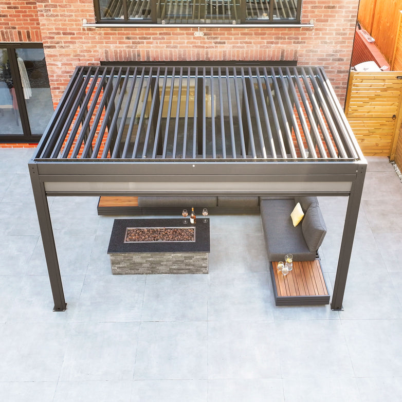 A modern outdoor lounge featuring a PergoSTET 3m x 4m Rectangular Pergola with 3 Drop Sides and LED Lighting in Grey, an L-shaped gray sofa, firepit, and wooden accents, all set against a brick wall. The space is thoughtfully designed with a built-in drainage system to handle any weather conditions effortlessly.