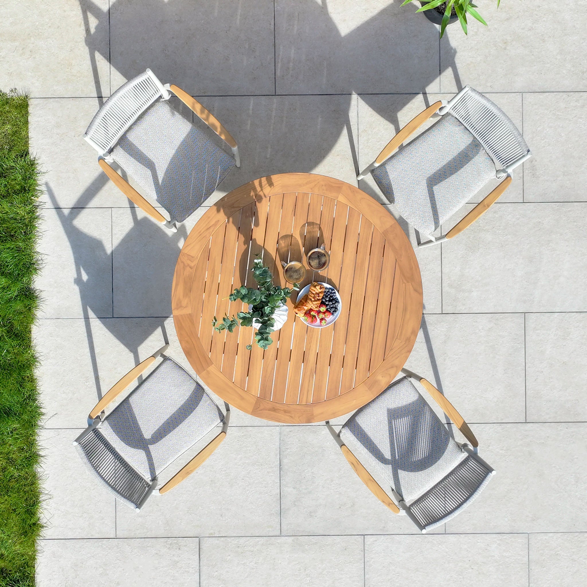 Top view of the Tellaro 4 Seat Round Dining Set in Latte, featuring a round teak table with drinks and a fruit bowl, accompanied by four outdoor chairs crafted from weather-resistant rope on a tiled terrace.