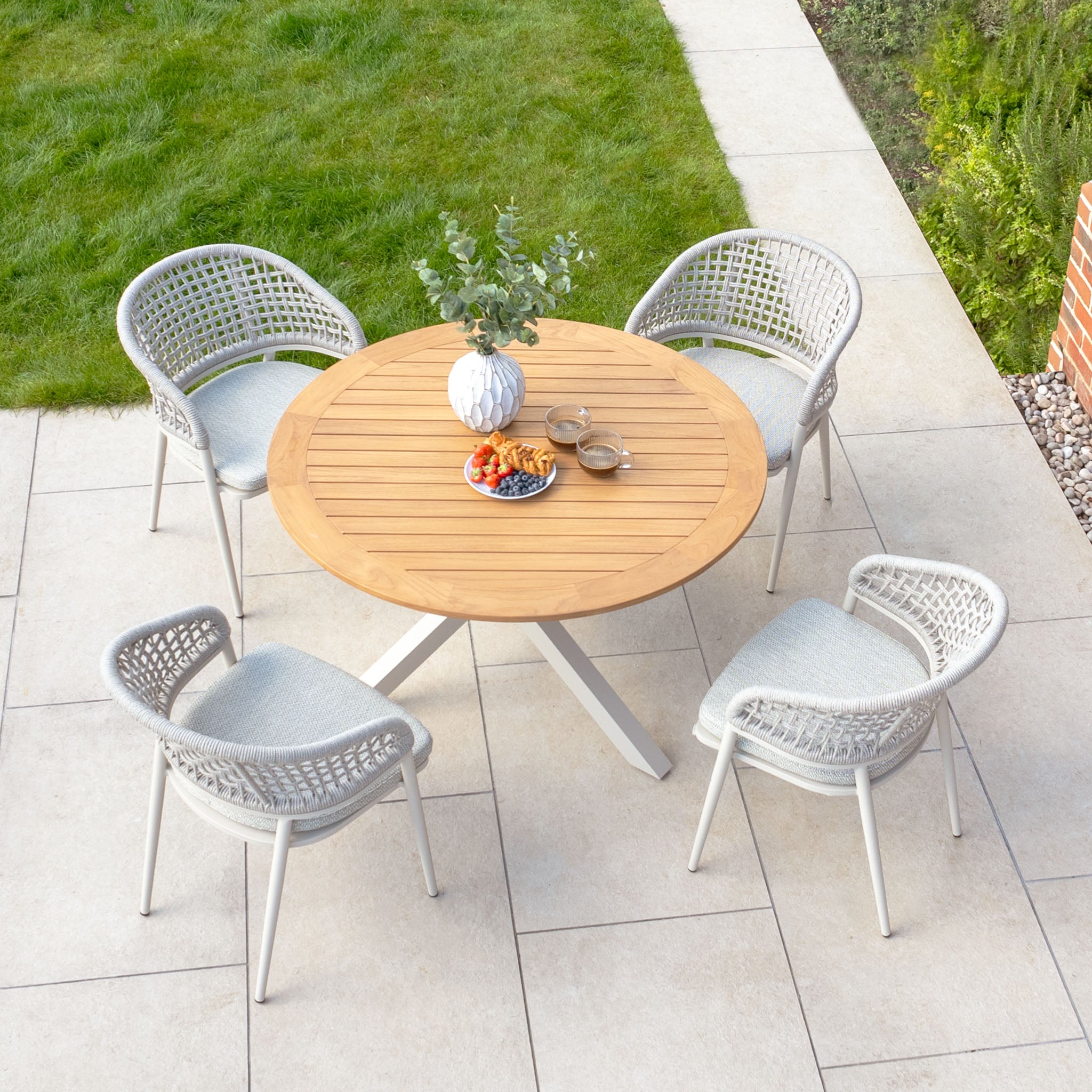 Outdoor patio showcasing the Kalama 4 Seat Round Dining Set in Latte, featuring a refined round teak table and four beautifully woven chairs, adorned with snacks, drinks, and a plant in a vase on the tabletop.