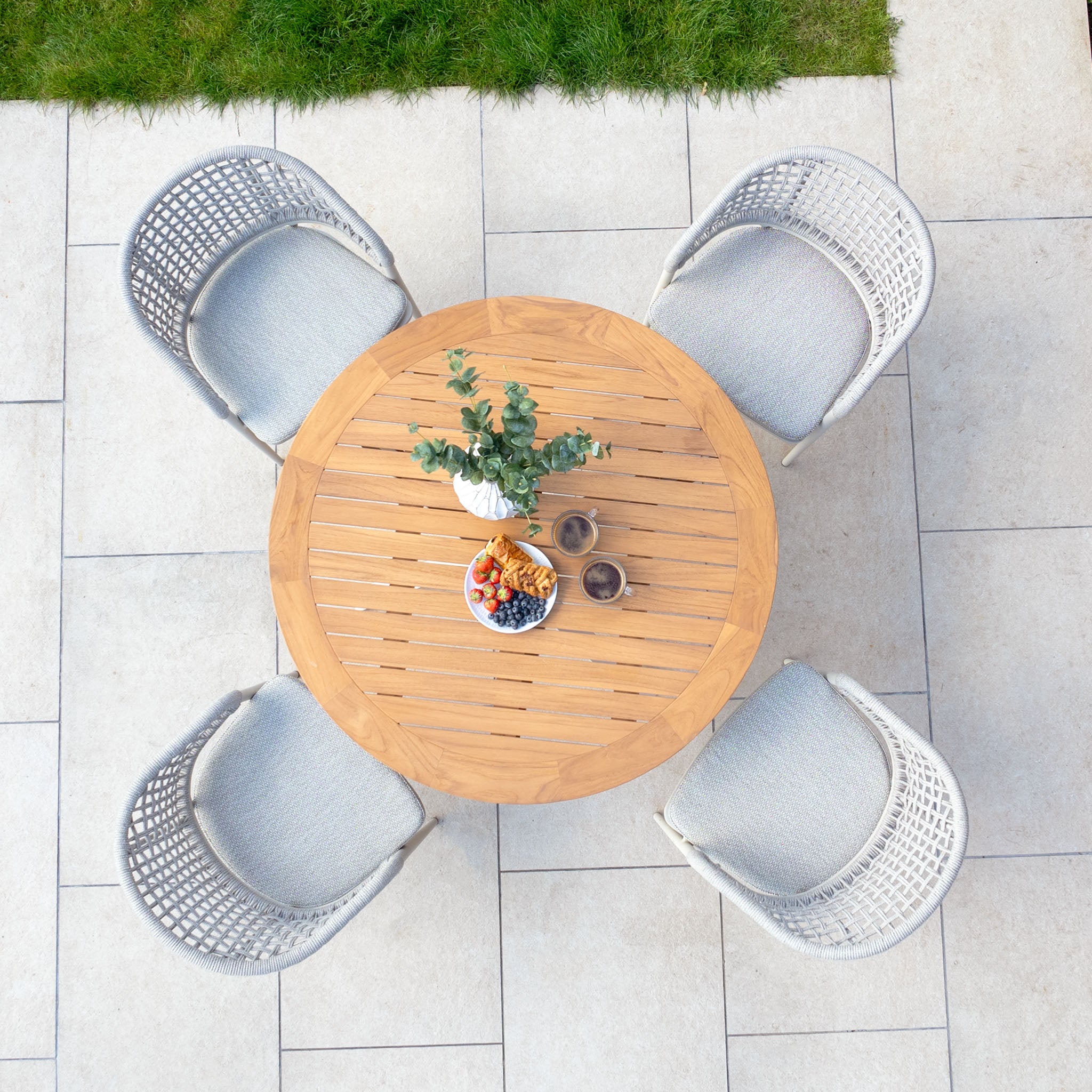 The Kalama 4 Seat Round Dining Set with a teak table in Latte, complemented by a plant and snacks, creates a cozy outdoor dining atmosphere with its woven chairs on the tiled patio.