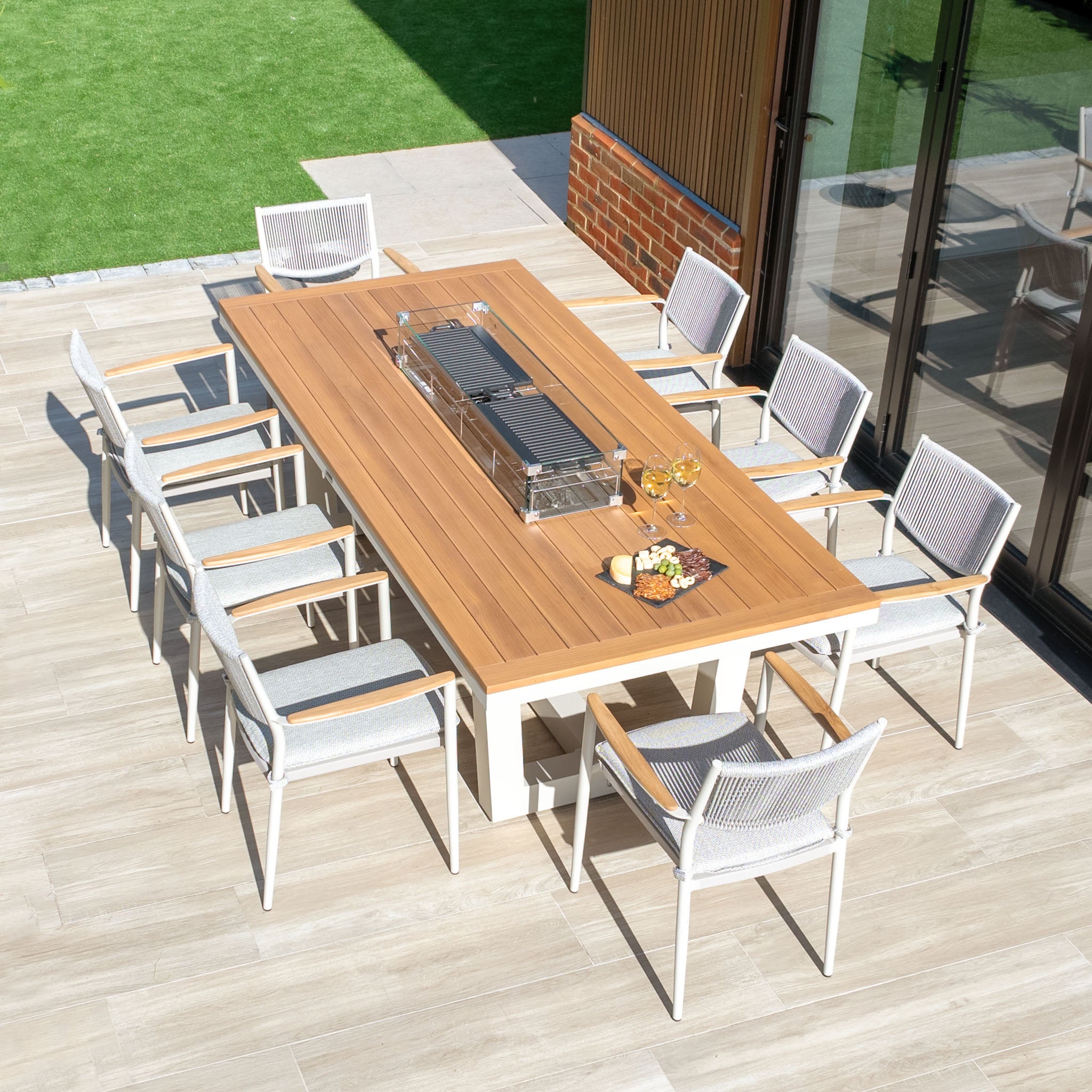 Outdoor dining set showcasing a spacious wooden table and a metal barbecue grill, complemented by Tellaro chairs on a patio next to the lawn. The arrangement features elegant powder-coated aluminium accents and effortlessly integrates with the Tellaro 8 Seat Rectangular Teak Firepit Dining Set in Latte for an elevated outdoor experience.