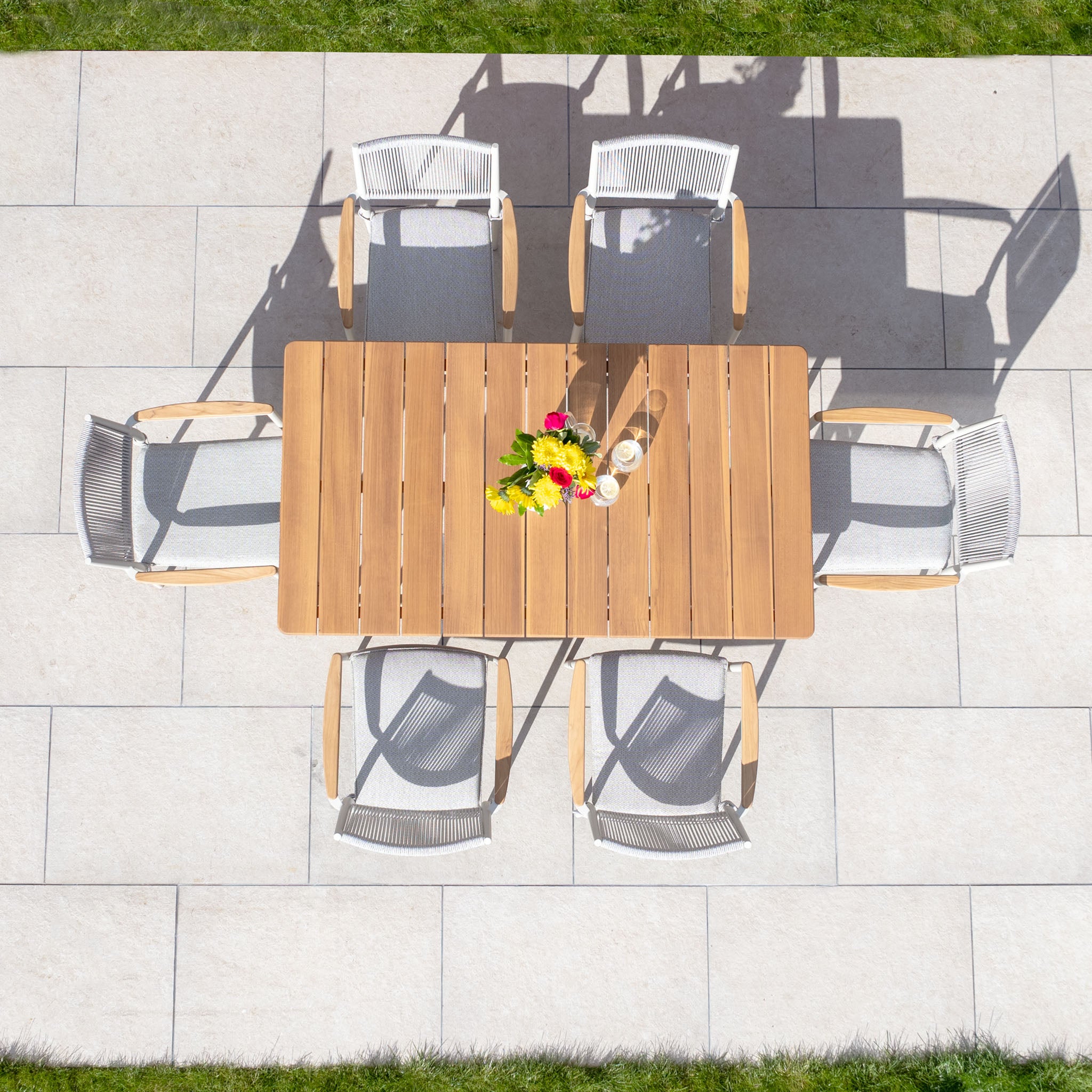 Top view of the Tellaro 6 Seat Rectangular Dining Set with Teak Table in Latte, featuring six chairs around a beautifully set table adorned with flowers and drinks on a stone patio. Experience outdoor living at its finest.