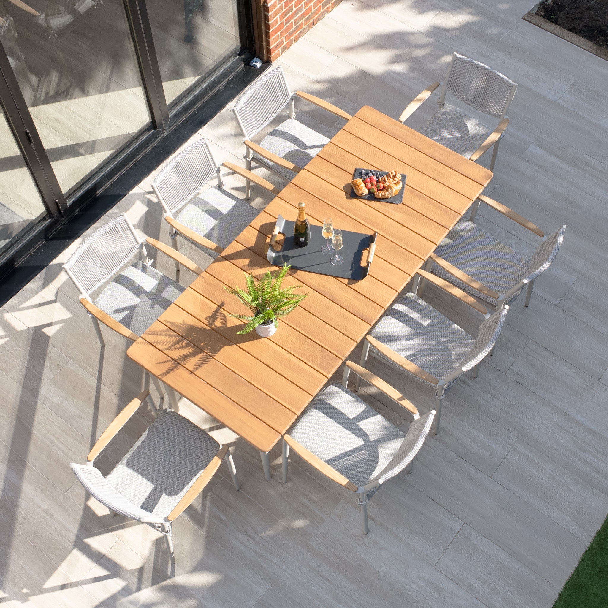 Enjoy outdoor dining with the Tellaro 8 Seat Rectangular Dining Set, featuring a weather-resistant teak table in Latte, complete with wine, glasses, and fruit under natural light. Ideal for alfresco occasions.