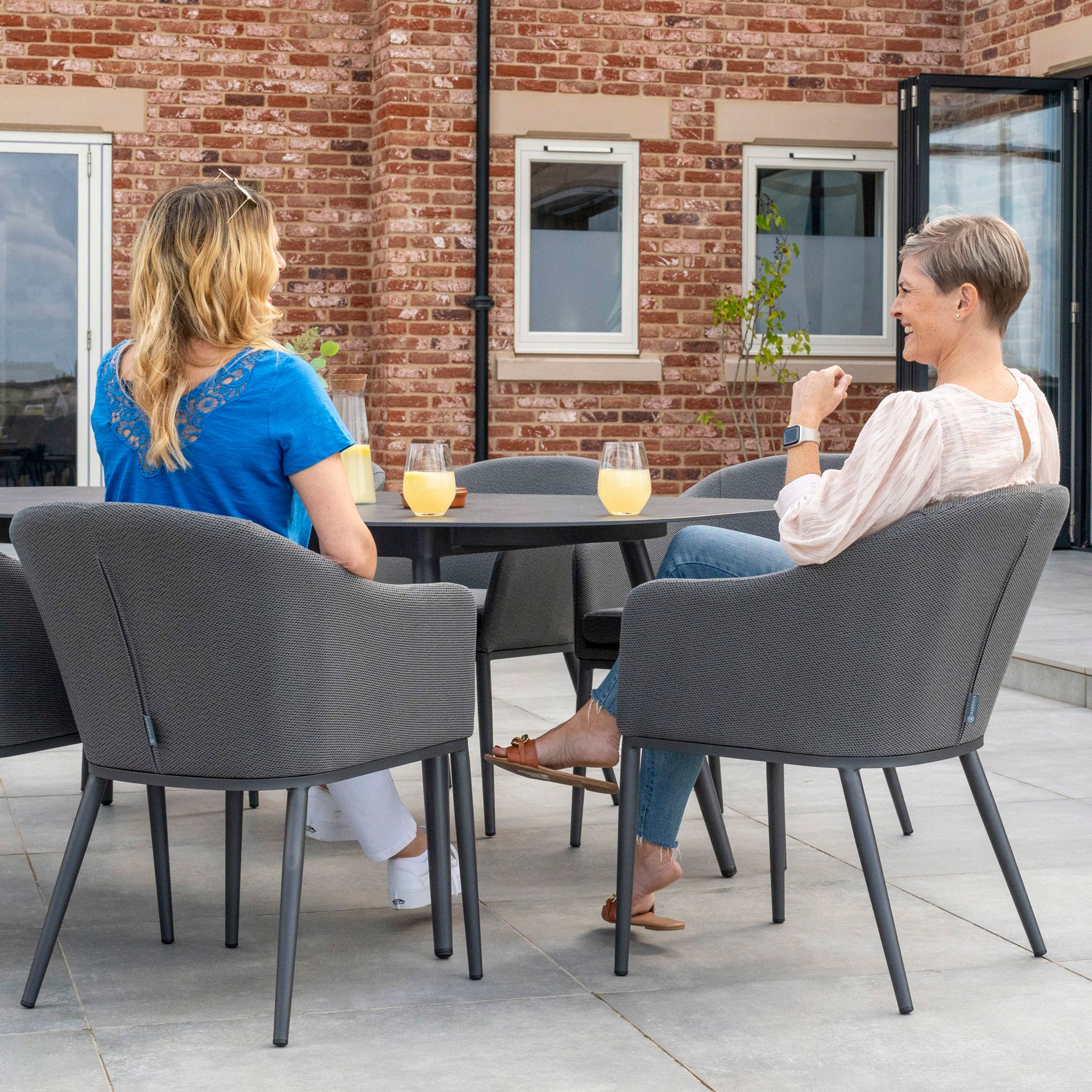 Two women sitting on gray chairs at a round table outside, enjoying their orange juice on an elegant Luna 6 Seat Outdoor Fabric Oval Ceramic Dining Set in Grey.