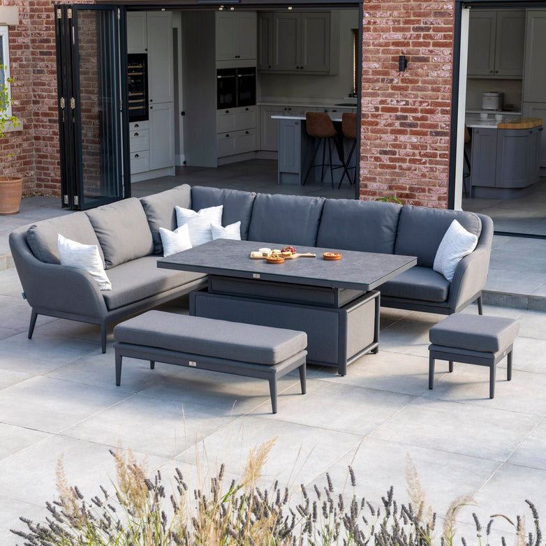 The Luna Outdoor Fabric Rectangular Corner Dining Set with Rising Table in Grey (Left Hand) from Harbour Lifestyle features a gray sectional sofa crafted with all-weather material, along with a table and benches. Set in a spacious backyard with a modern kitchen visible in the background, it's perfect for enjoying sunny days and cozy evenings alike.