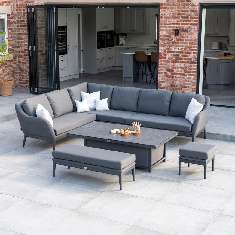 Outdoor patio with the Luna Outdoor Fabric Rectangular Corner Dining Set featuring a rising table in grey (left hand), complemented by pillows, coffee table, and ottomans on tiled flooring; brick house in the background. Enjoy the Harbour Lifestyle with durable all-weather material designed to withstand the elements.