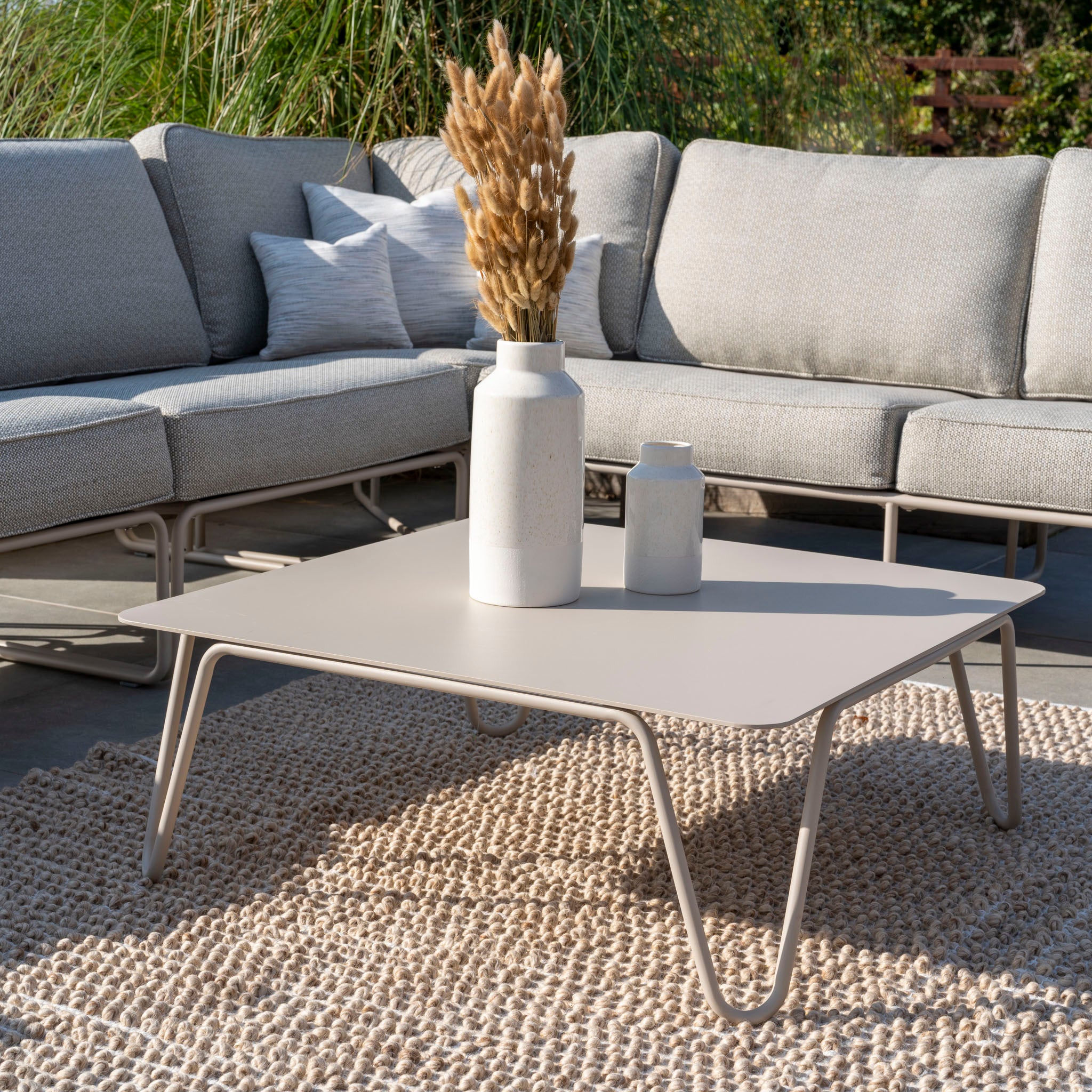 Experience the Riviera Luxury Large Outdoor Corner Group Set in Latte, featuring a gray sectional sofa, beige table, teak armrests, decorative vases, and a woven rug. The hydrophobic rope weave ensures lasting comfort and style in any weather.