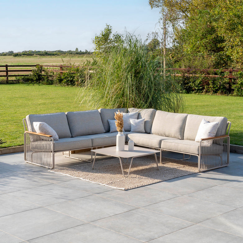 Outdoor patio with a beige sectional sofa and table on a rug, surrounded by lush greenery and a wooden fence, part of the Riviera Luxury Outdoor Corner Group Set in Latte featuring hydrophobic rope weave and Rust Proof Tiger Drylac Powder Coating.