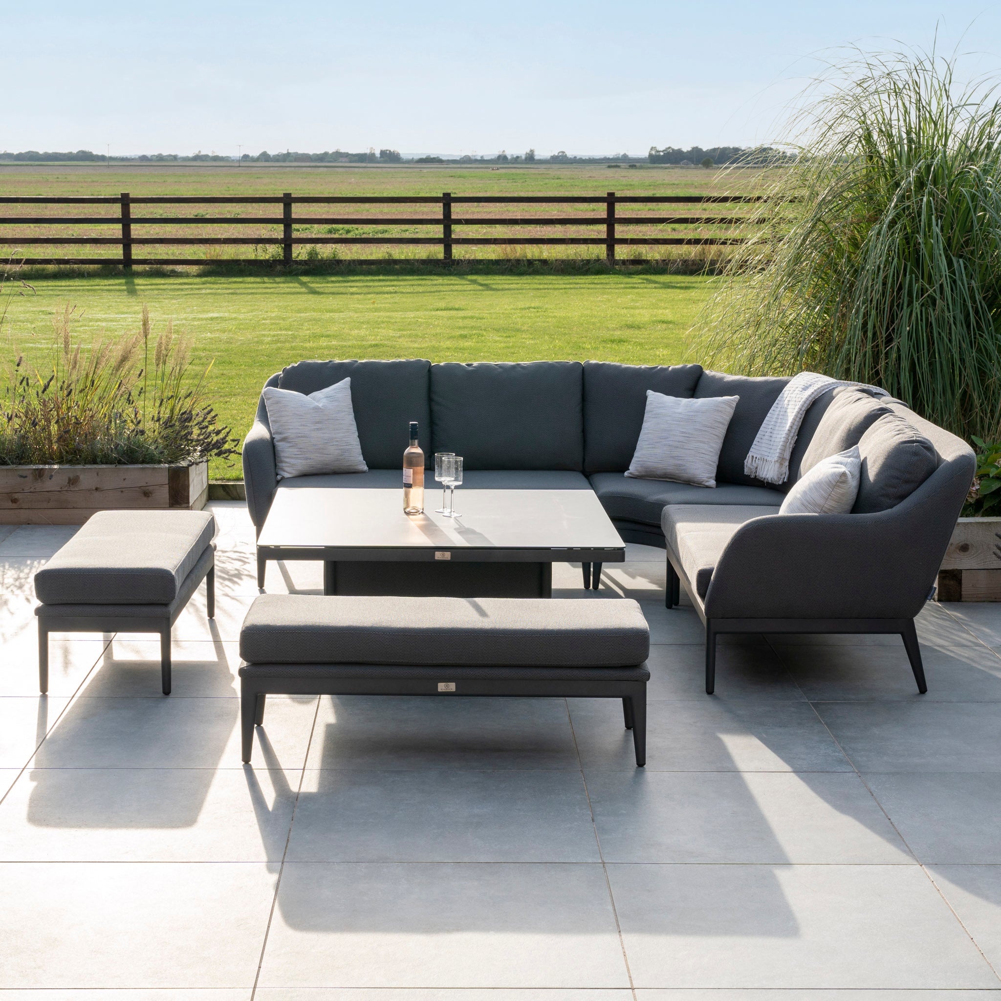 Outdoor patio set featuring the Luna Deluxe Outdoor Fabric Square Corner Dining Set with Rising Table in Grey, arranged on a tiled patio overlooking a green field.