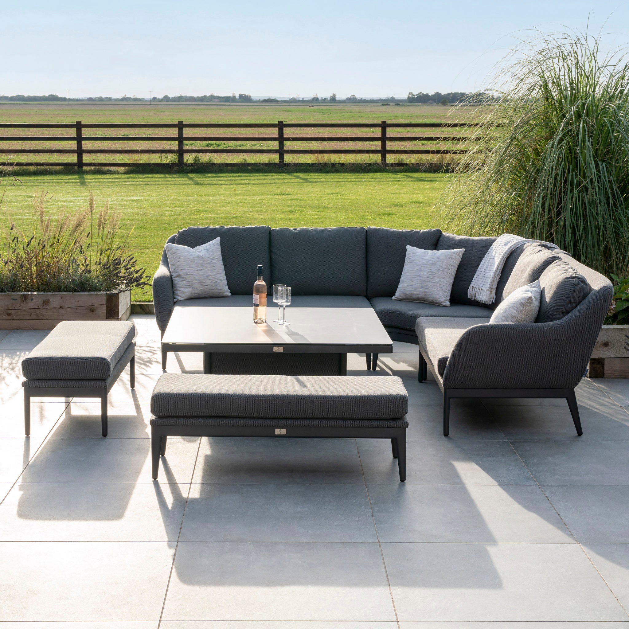 Outdoor patio with a Harbour Lifestyle Luna Deluxe Outdoor Fabric Square Corner Dining Set in Grey, featuring an all-weather material sectional sofa, a rising table with a bottle of wine and glasses, two benches, and scenic field views.