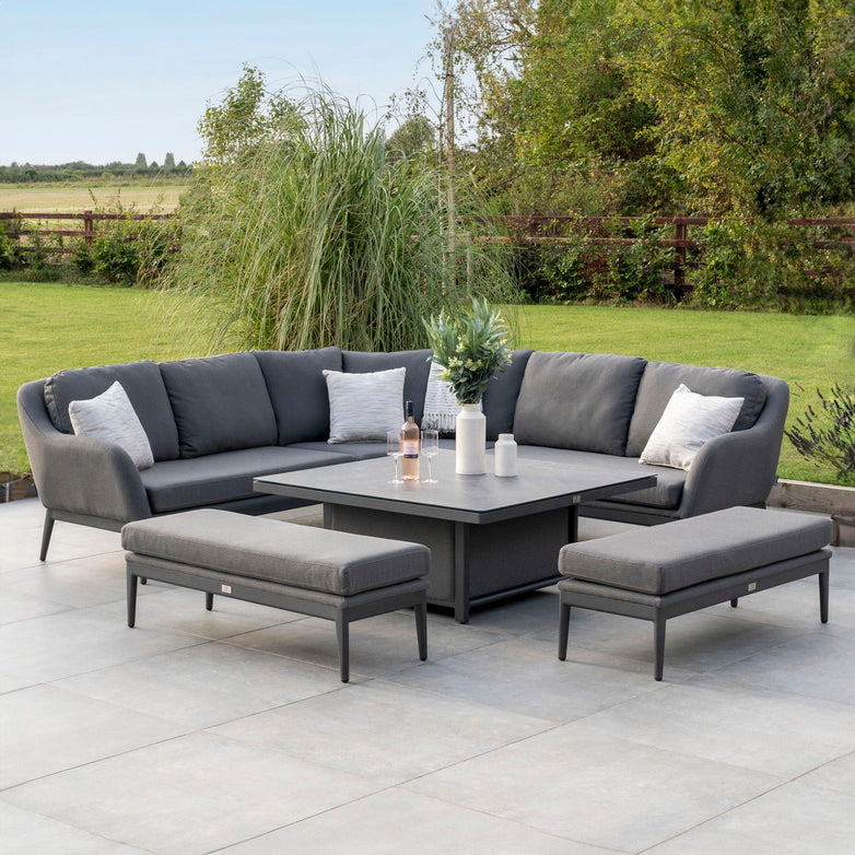 Modern outdoor patio with a gray sectional sofa, matching benches, and a central table, all featuring the Harbour Lifestyle Luna Deluxe Outdoor Fabric Square Corner Dining Set with Rising Table in Grey. The setup is set against a lush green backdrop for a perfect blend of style and durability using all-weather material.