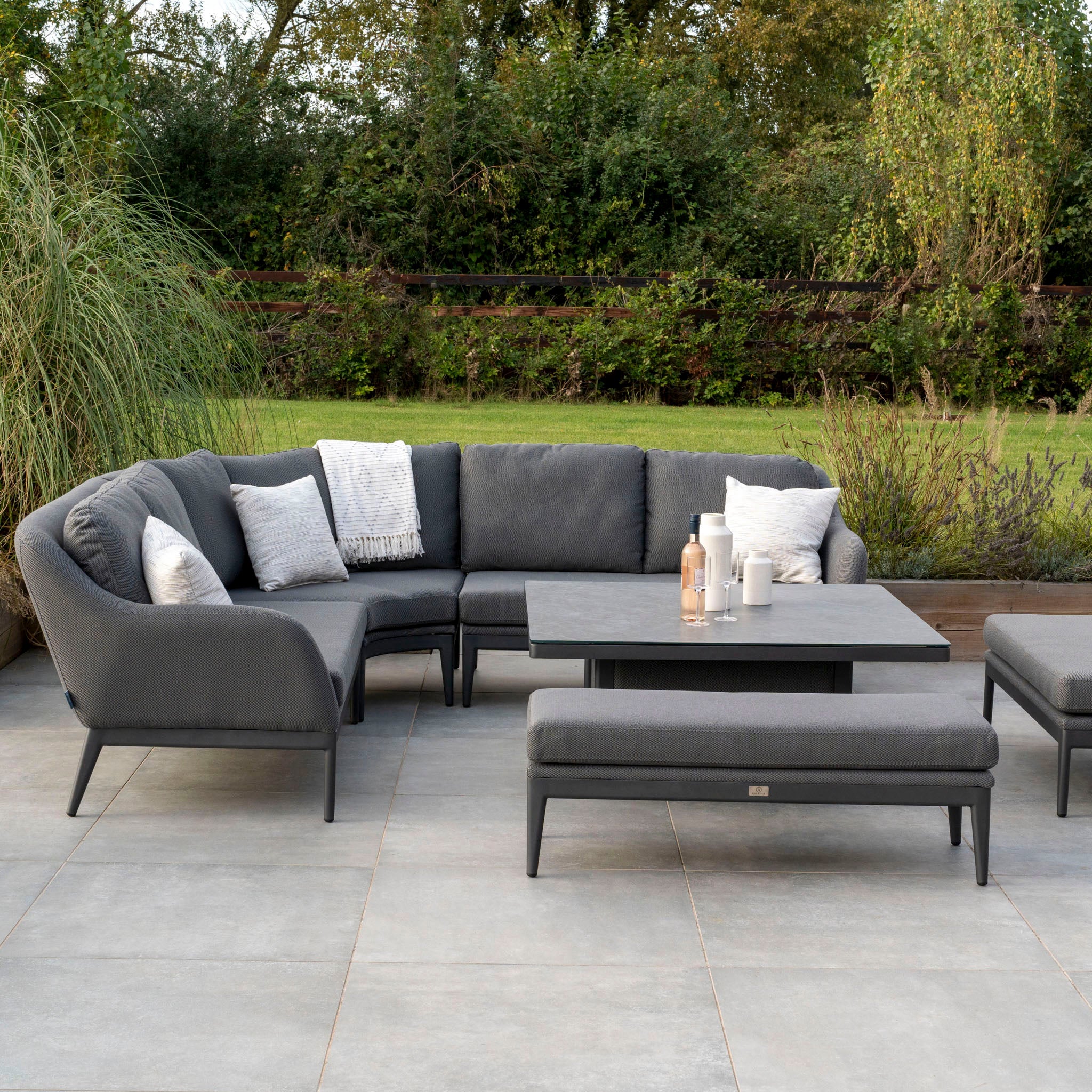 Outdoor patio with a gray sectional sofa, pillows, matching ottoman, and a coffee table. Crafted from all-weather material, this setup features the Harbour Lifestyle Luna Deluxe Outdoor Fabric Square Corner Dining Set with Rising Table in Grey. Green trees provide a serene background.