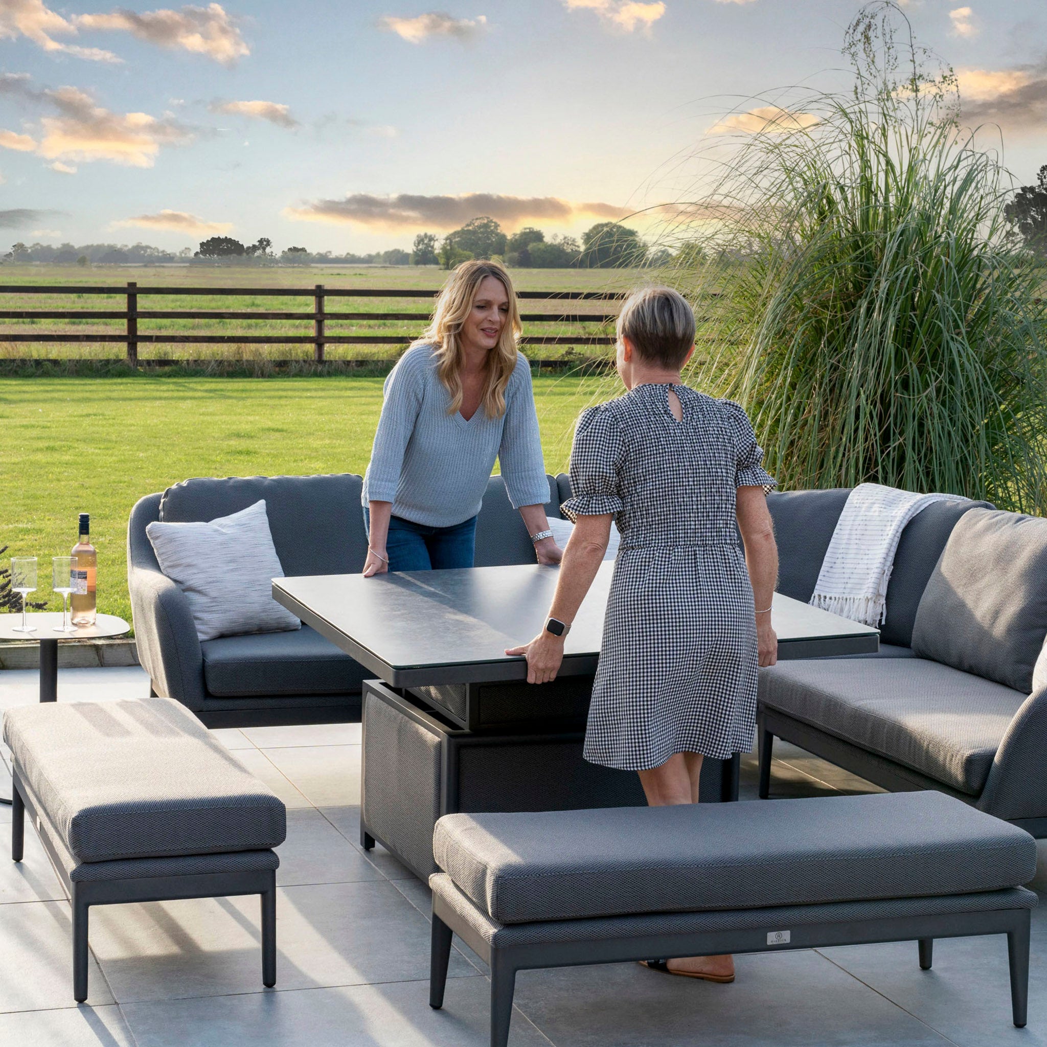 Two women adjusting a Harbour Lifestyle Luna Deluxe Outdoor Fabric Square Corner Dining Set with Rising Table in Grey on a patio with outdoor seating, a bottle of wine, and a scenic countryside backdrop at sunset.