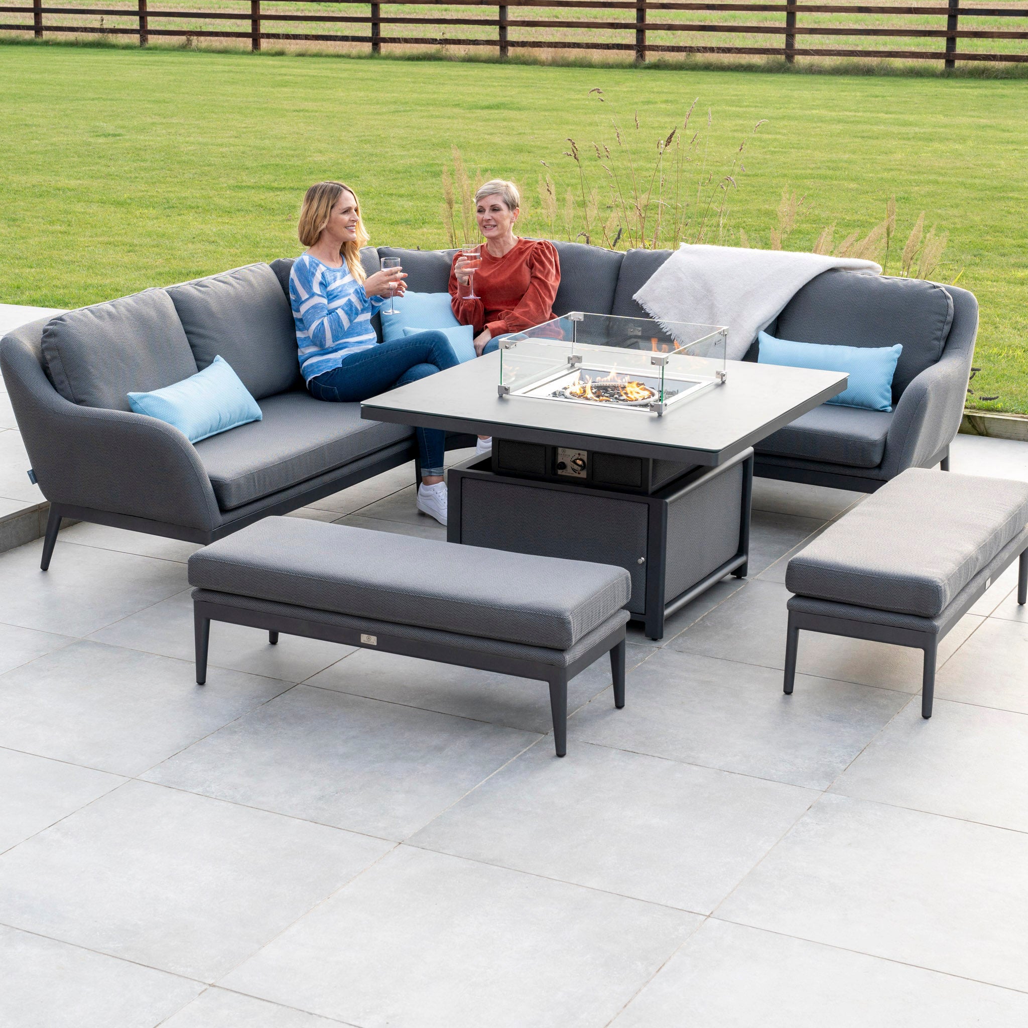 Two people sitting on a Luna Deluxe Outdoor Fabric Square Corner Dining Set with Rising Firepit Table in Grey, enjoying drinks near the adjustable firepit table on a spacious patio. The setup is both stylish and maintenance-free.