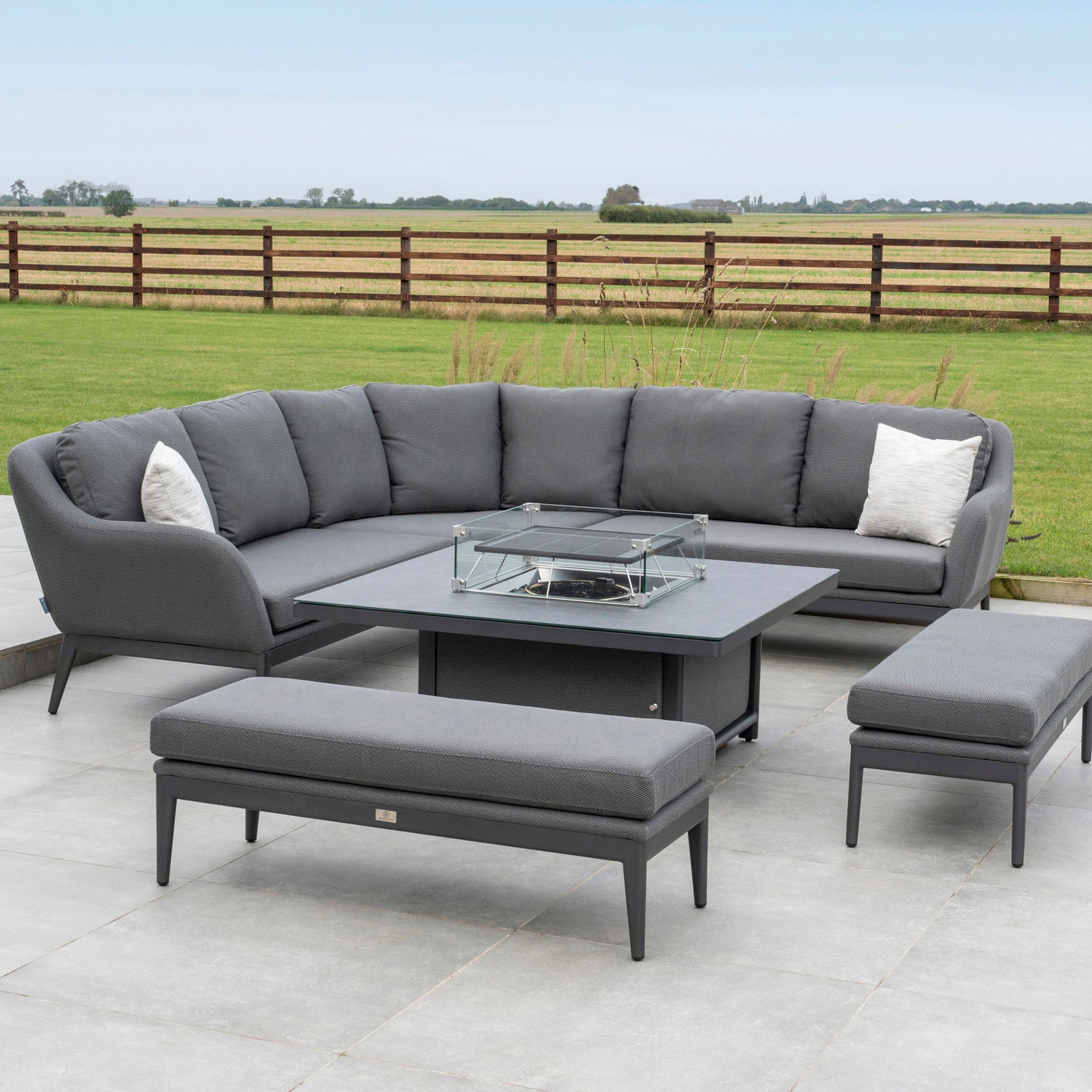 Luna Deluxe Outdoor Fabric Square Corner Dining Set with a Rising Firepit Table in Grey, featuring maintenance-free fabric, a corner sofa, benches, and set against a vast open field background.