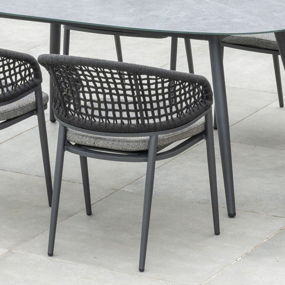 The Kalama 6 Seat Rope Oval Dining Set with Ceramic Table in Charcoal boasts a weather-proof aluminum frame and stylish woven rope chairs, all elegantly arranged around a sleek ceramic glass table on a tiled surface.