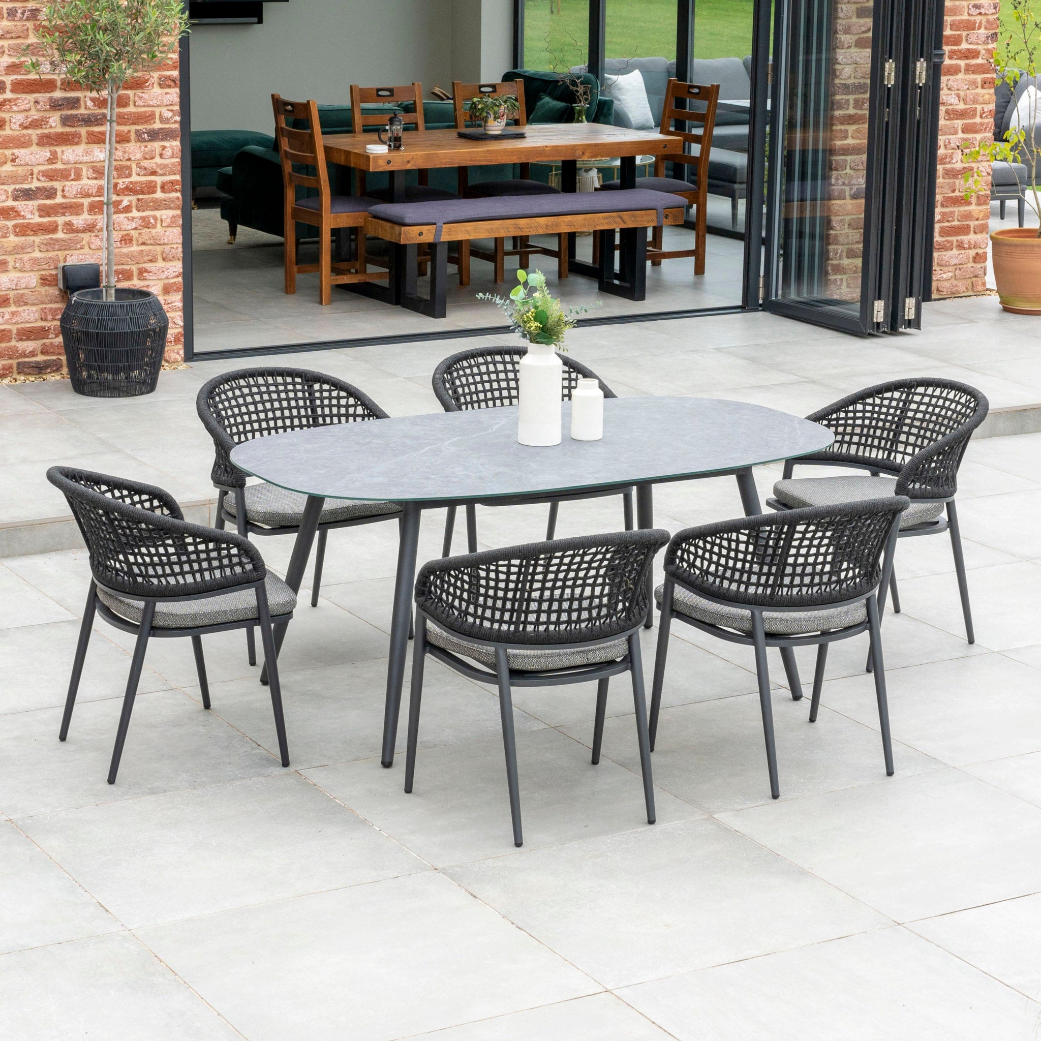 Outdoor patio with the Kalama 6 Seat Rope Oval Dining Set featuring a ceramic table in charcoal, adjacent to an indoor dining room with wooden furniture.