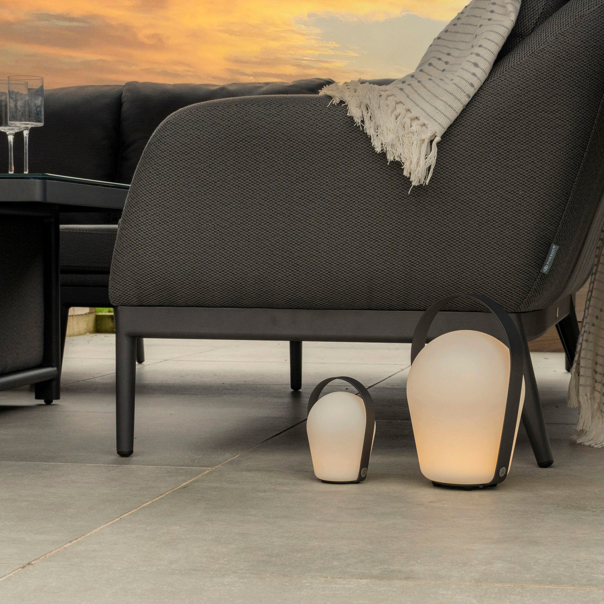 Three Bob Small Table Lights in Charcoal near a gray chaise lounge with a blanket, set on a patio during sunset.