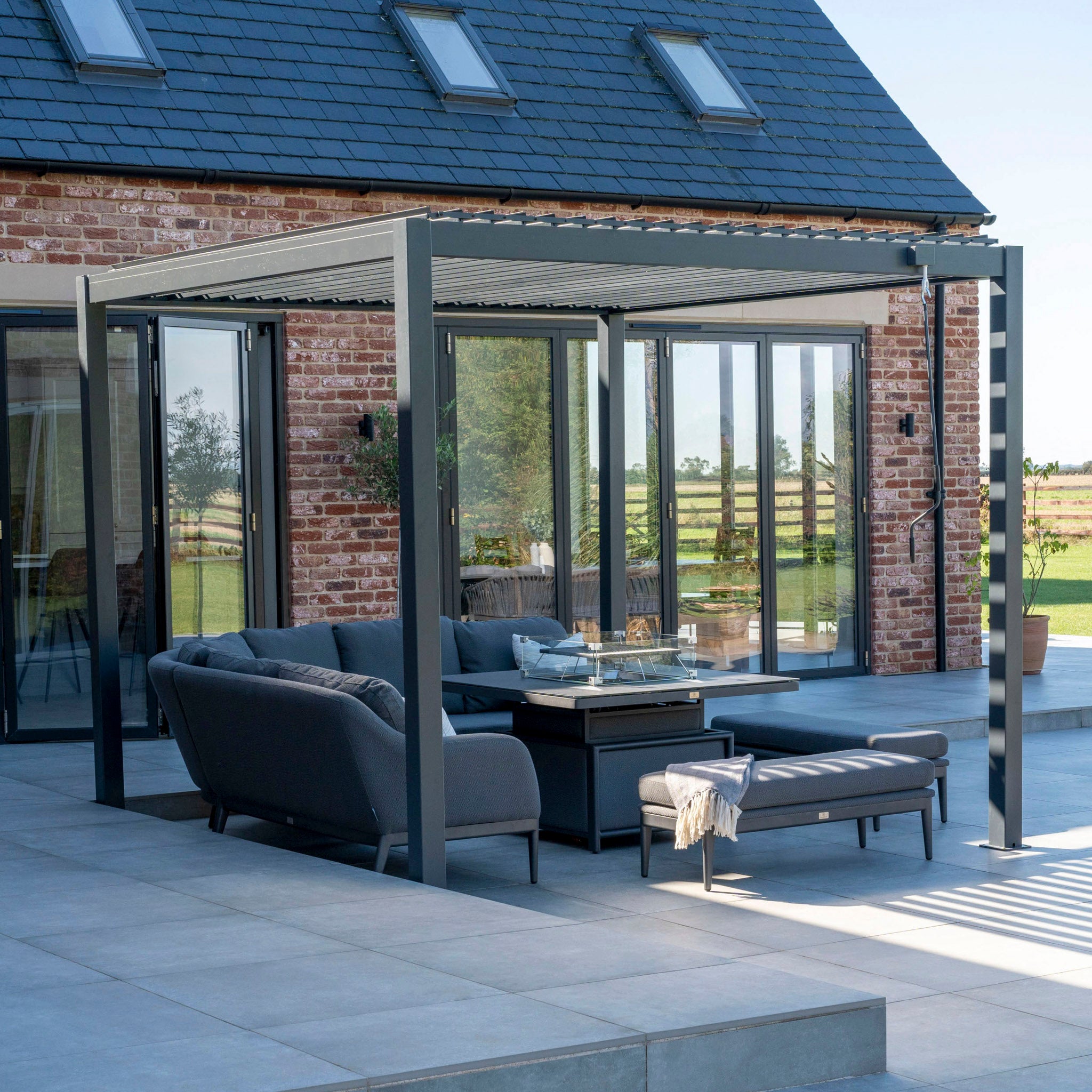 Modern outdoor patio featuring the Luna Deluxe Outdoor Fabric Square Corner Dining Set with a Rising Firepit Table in Grey under a pergola, adjacent to a brick house with large glass doors. The maintenance-free fabric of the grey sofas and the firepit table complete the cozy yet stylish setup.